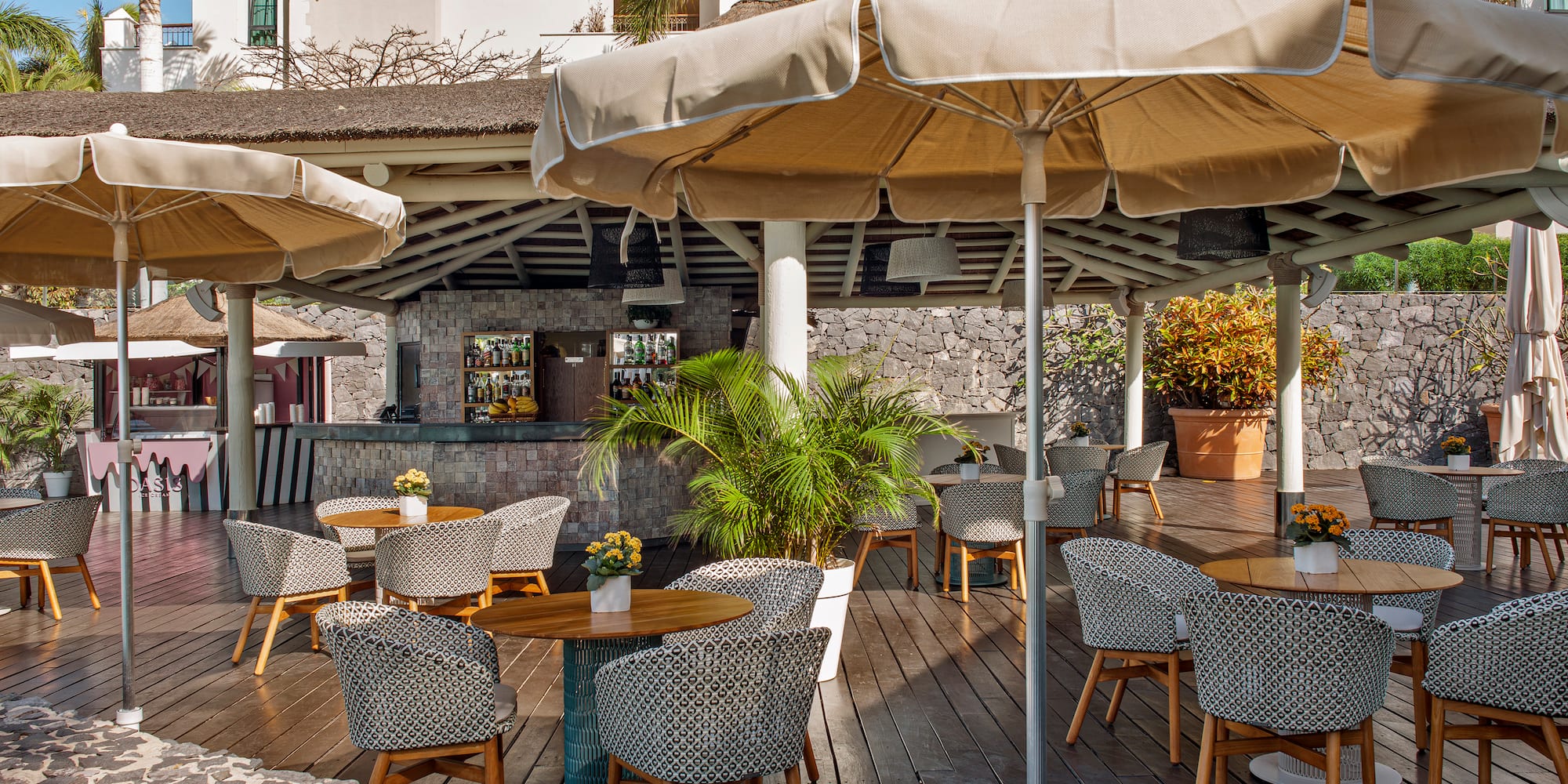 Tropical outdoor restaurant patio with bar, shaded tables, and patterned chairs.