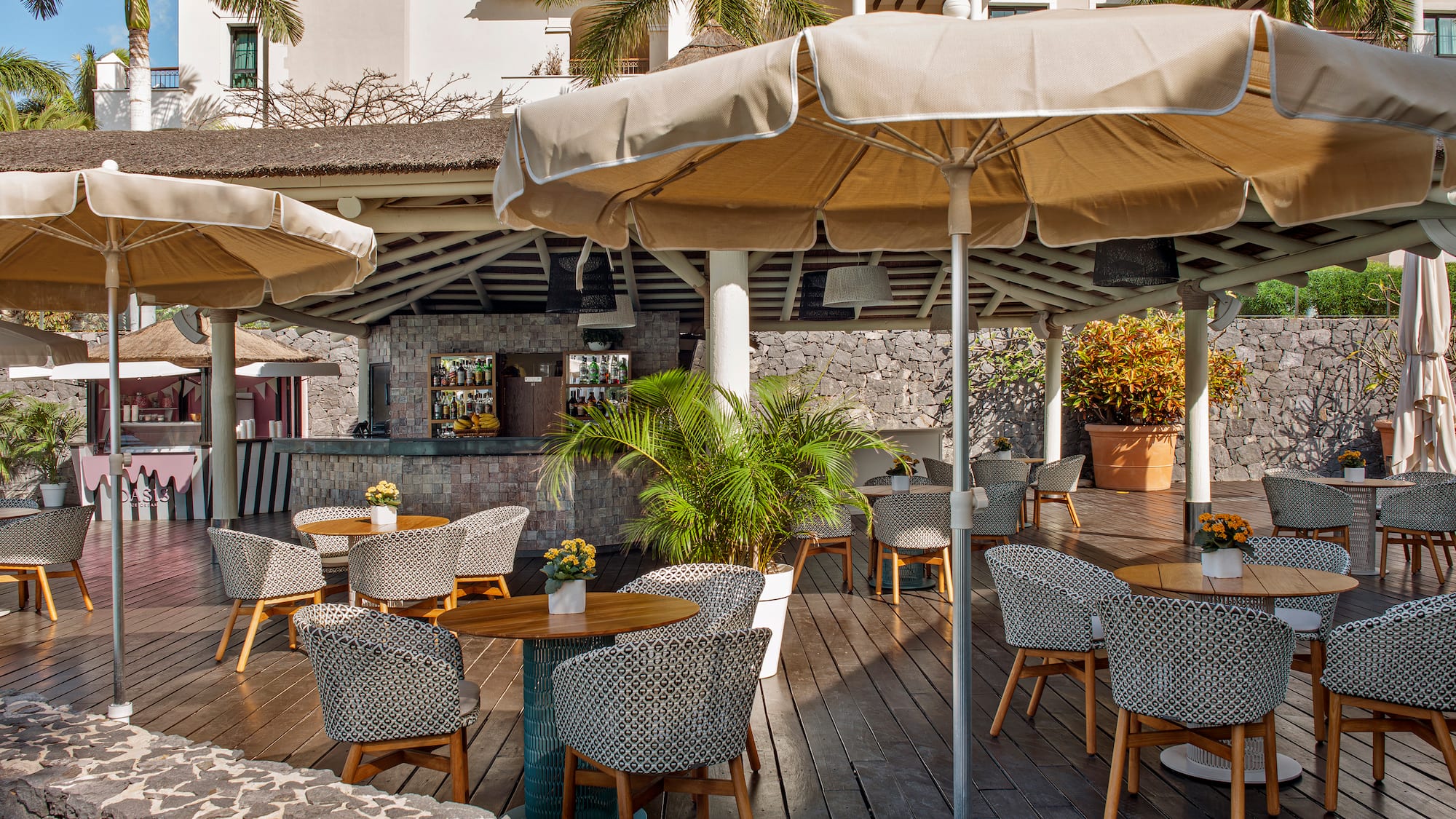 Tropical outdoor restaurant patio with bar, shaded tables, and patterned chairs.