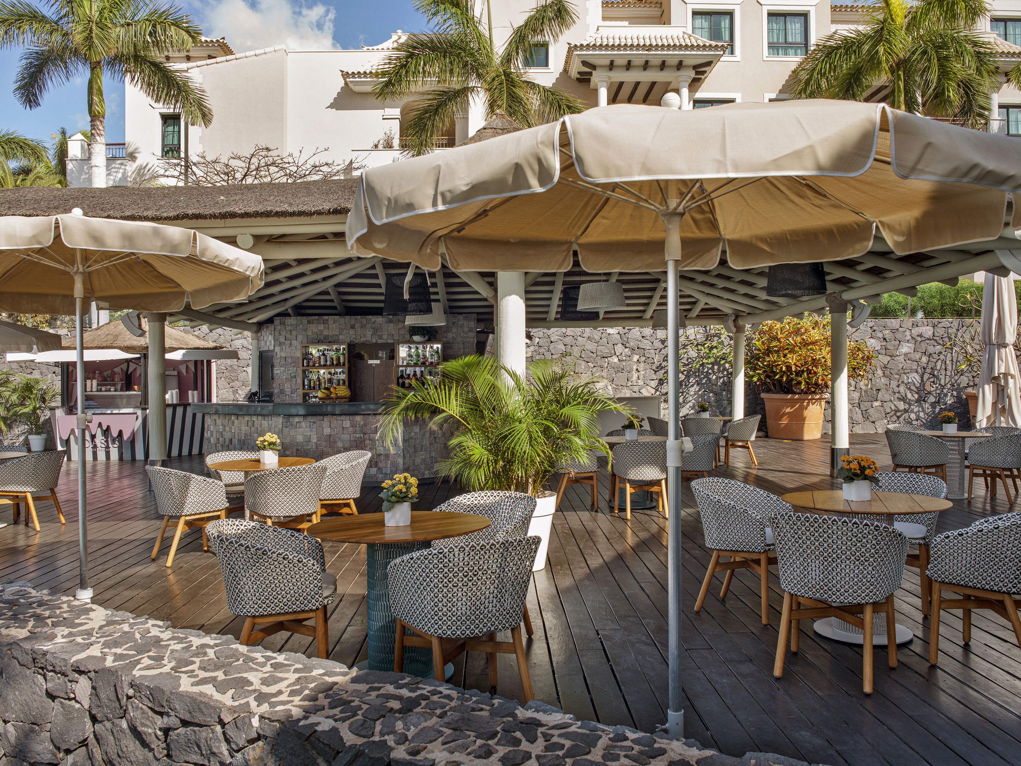 Tropical outdoor restaurant patio with bar, shaded tables, and patterned chairs.