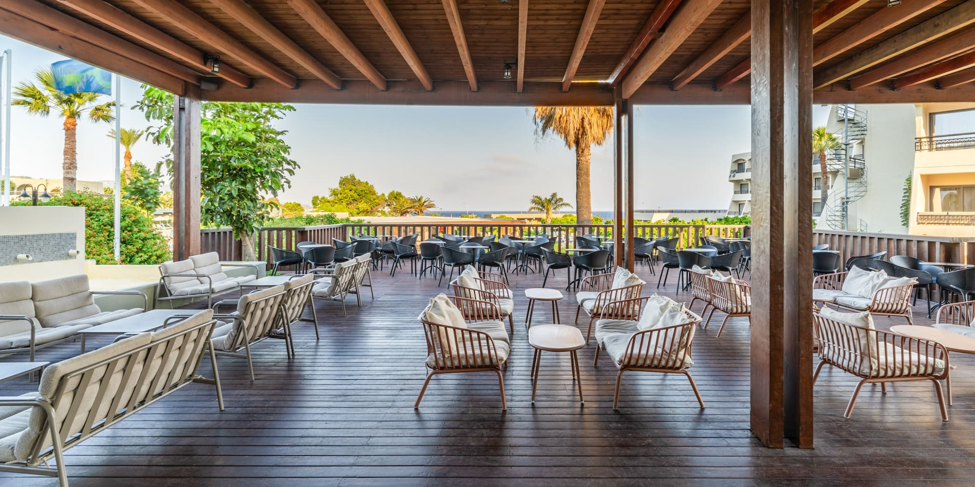 Covered resort patio with lounge seating, wooden deck, palm trees, sea view.