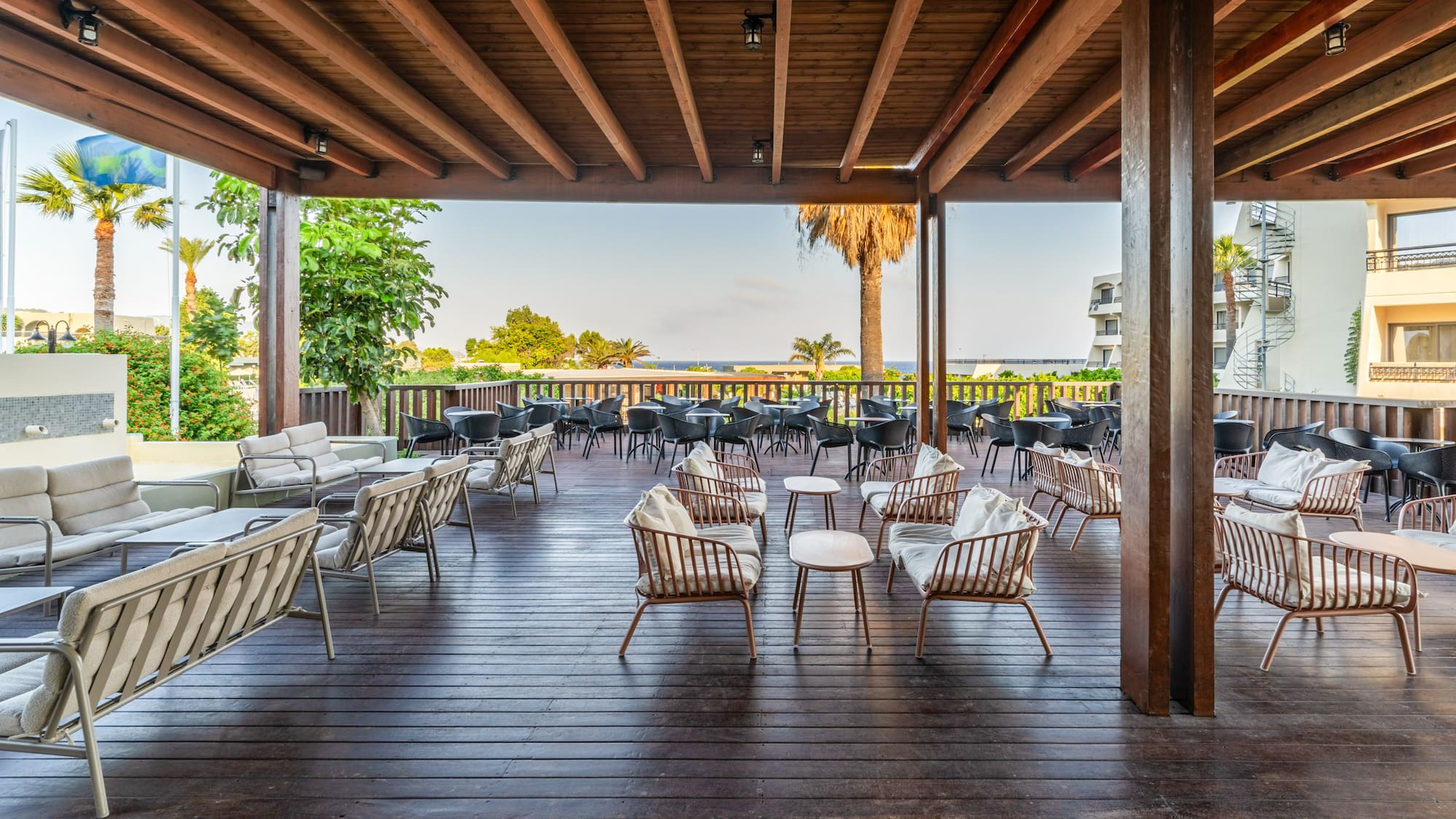 Covered resort patio with lounge seating, wooden deck, palm trees, sea view.