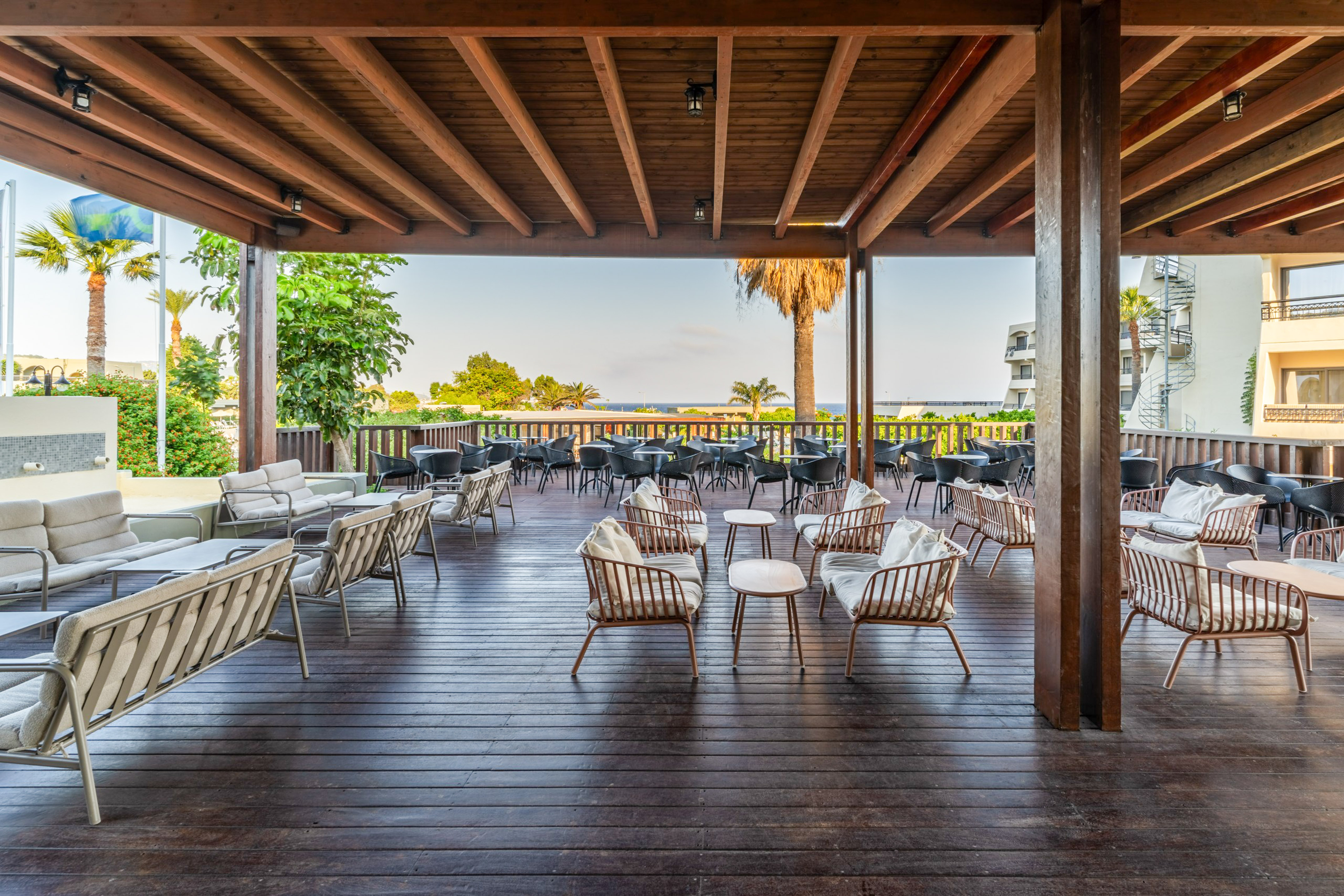 Covered resort patio with lounge seating, wooden deck, palm trees, sea view.