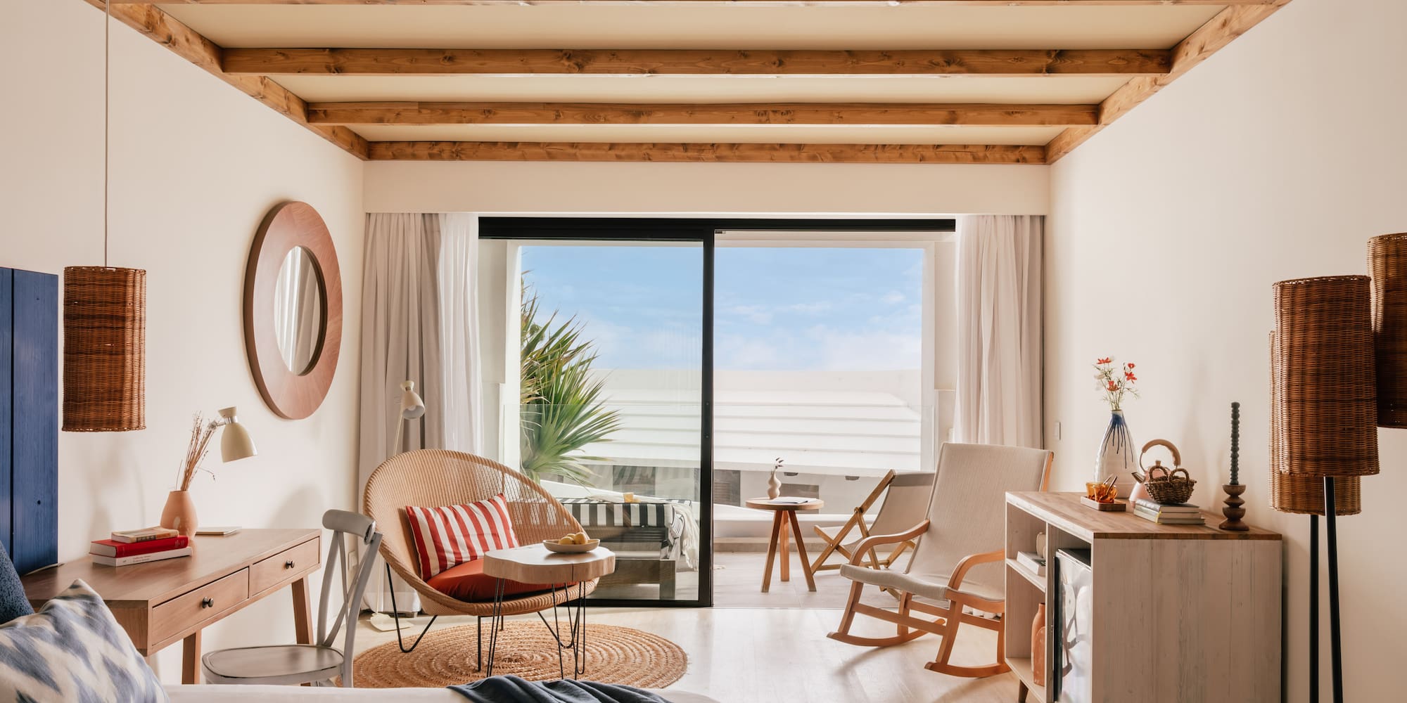 Habitación de hotel con balcón, cama, silla de mimbre y techo de vigas.