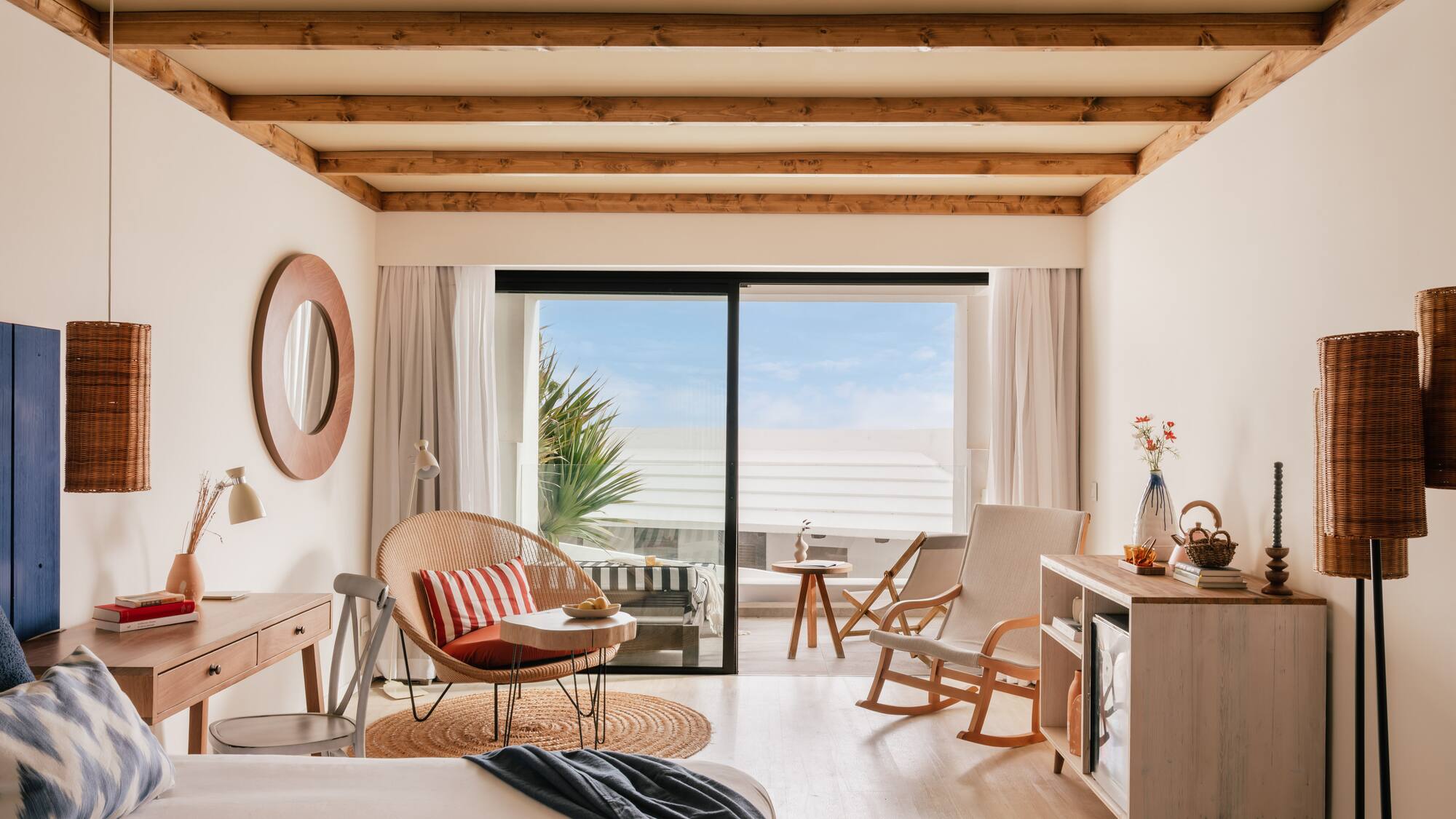 Habitación de hotel con balcón, cama, silla de mimbre y techo de vigas.