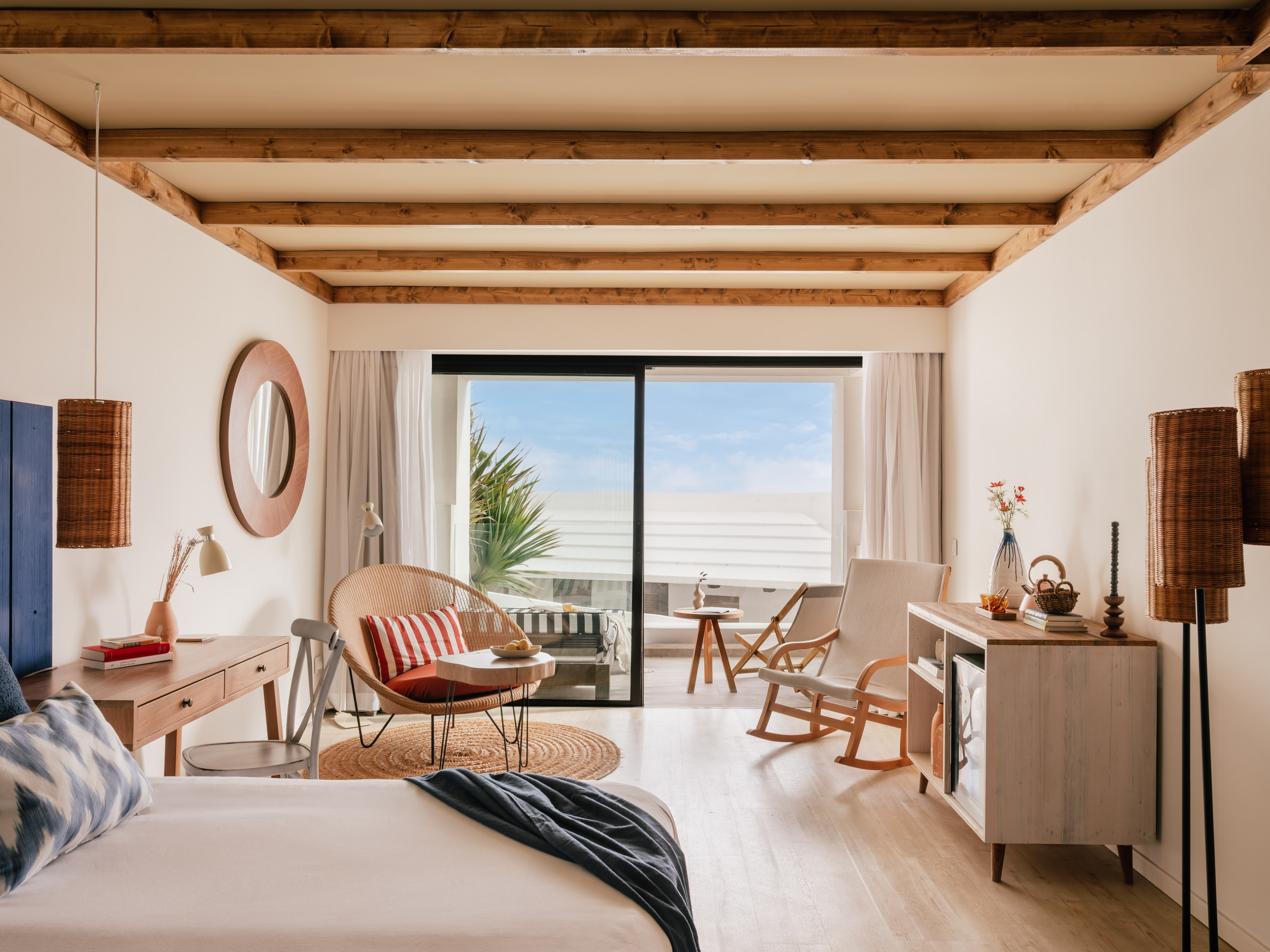 Habitación de hotel con balcón, cama, silla de mimbre y techo de vigas.