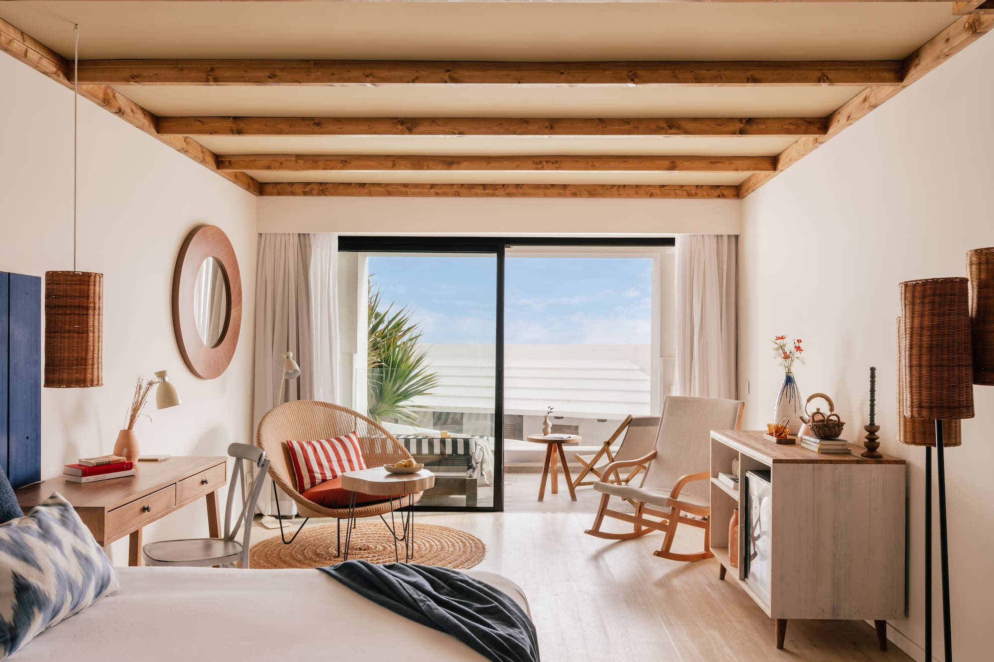 Habitación de hotel con balcón, cama, silla de mimbre y techo de vigas.