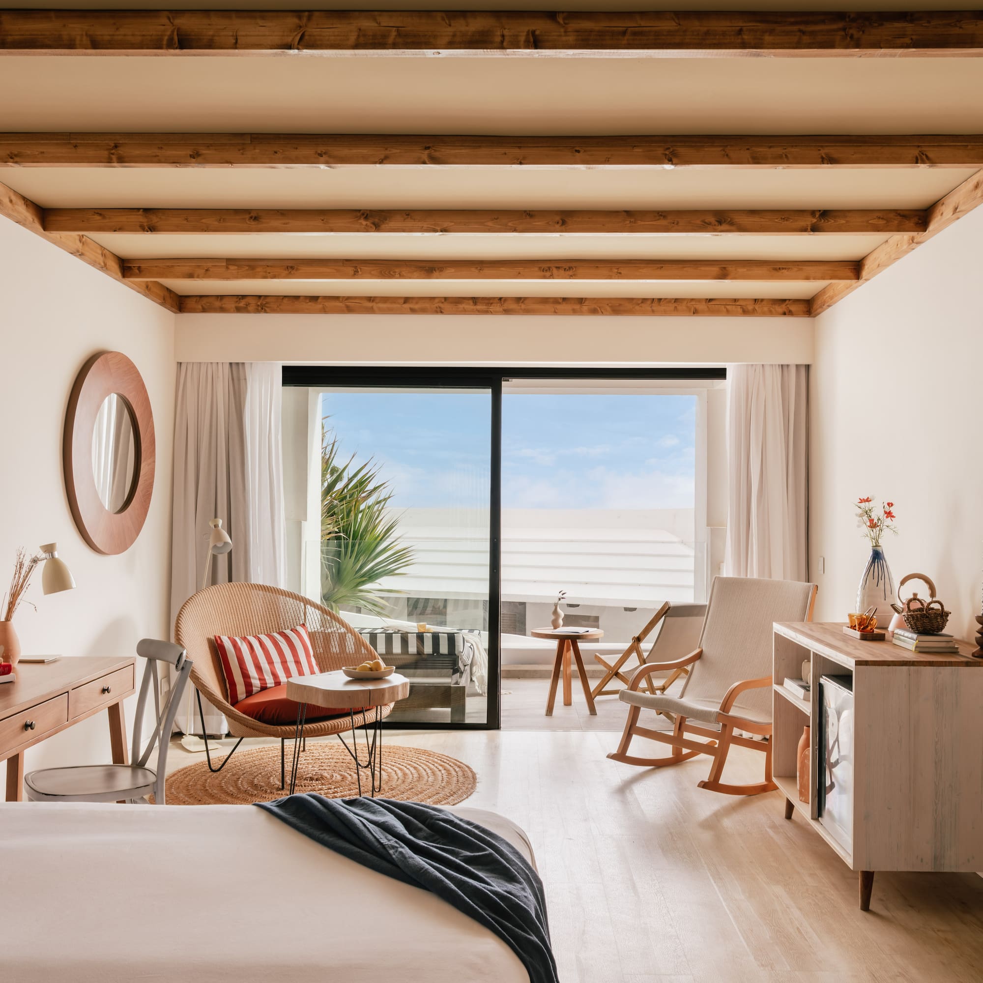 Habitación de hotel con balcón, cama, silla de mimbre y techo de vigas.