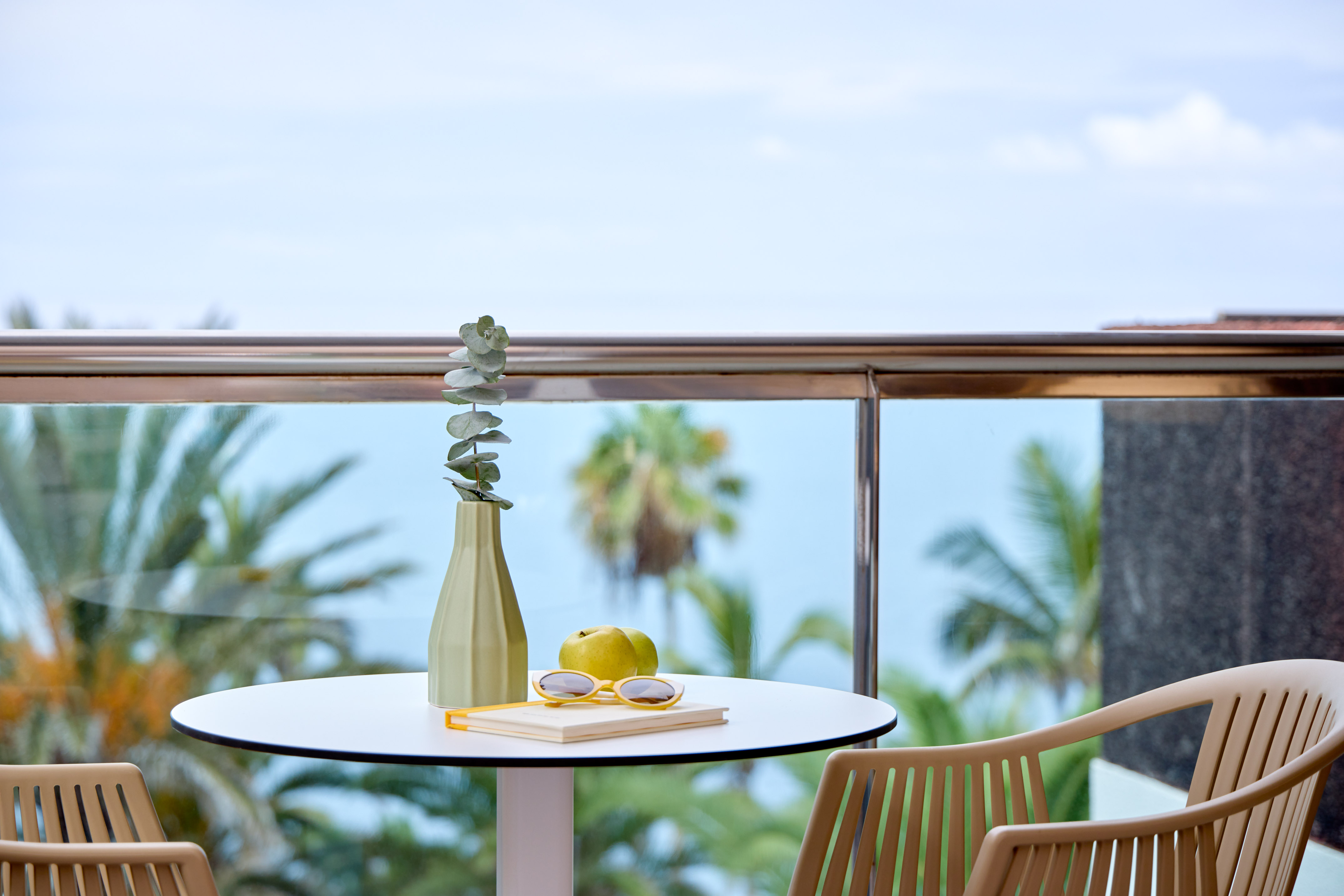 Balcony table with eucalyptus, apples, sunglasses, and tropical palm trees.