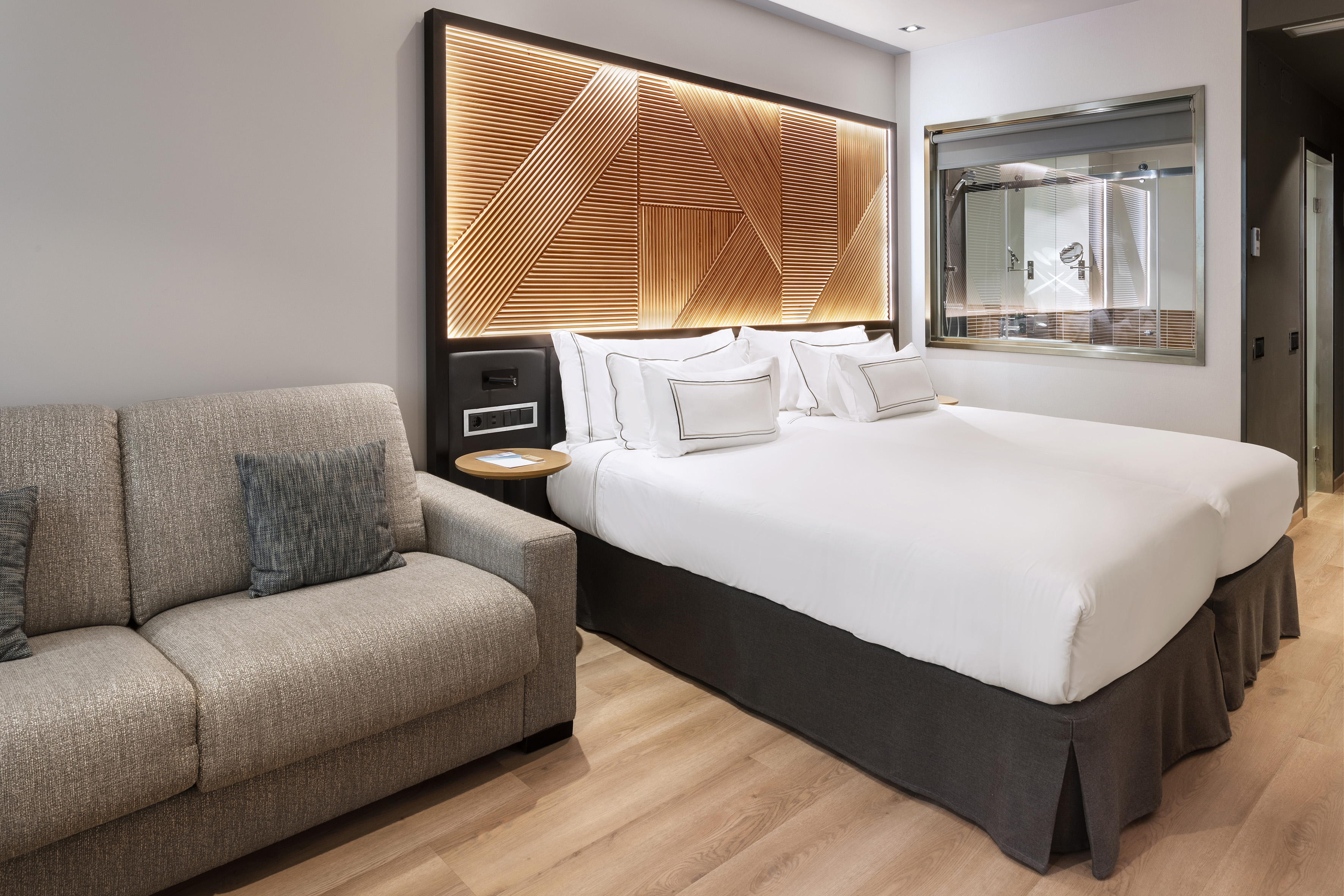 Modern hotel bedroom with white bed, backlit wooden headboard, and grey sofa.
