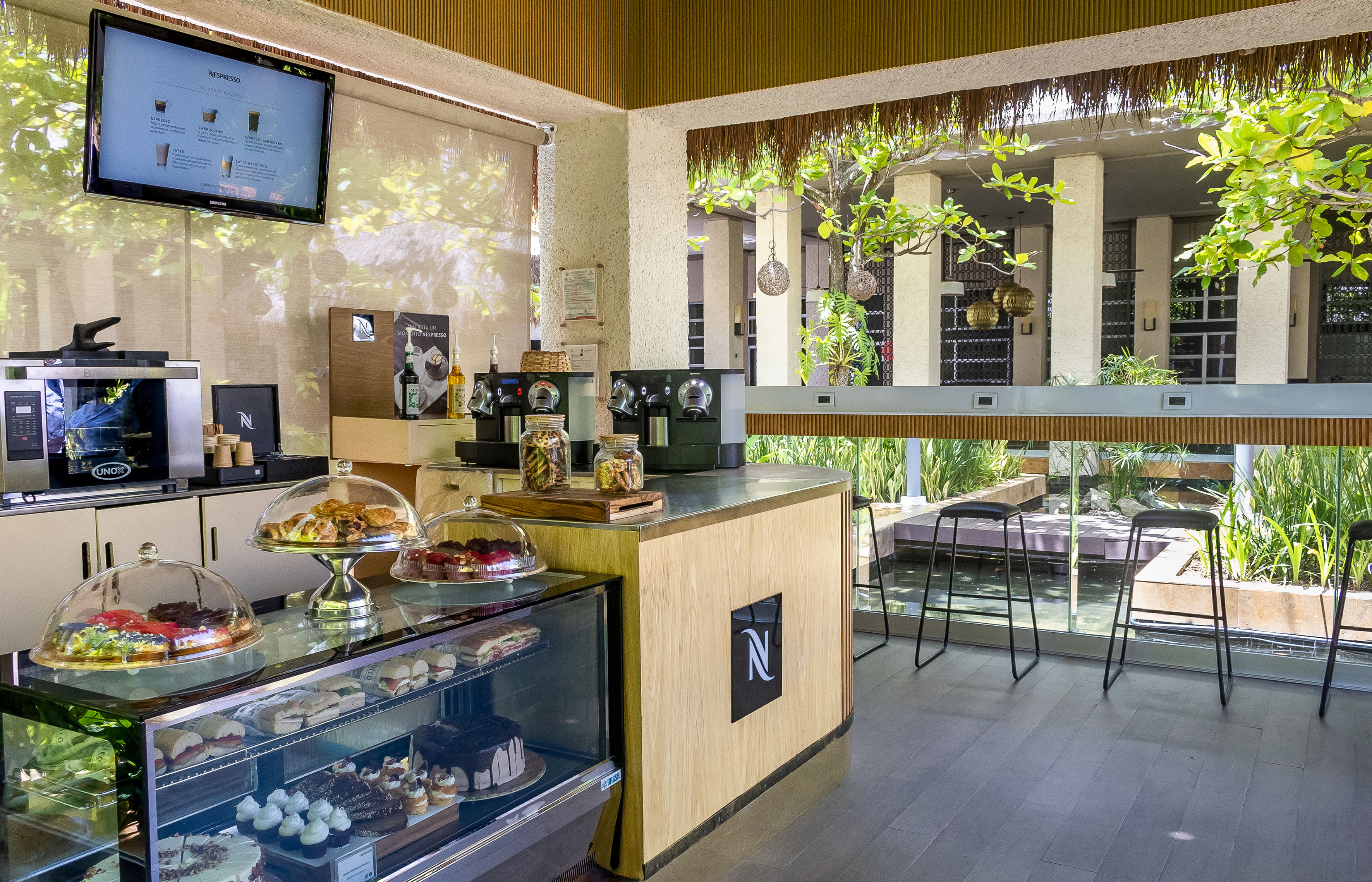Modern Nespresso coffee bar with pastries, cakes, and tropical outdoor view.