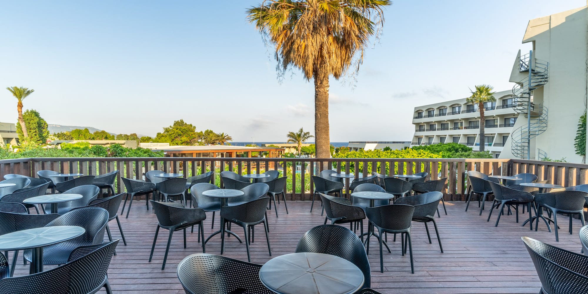 Sunny hotel patio with black outdoor seating, palm tree, and distant sea view.