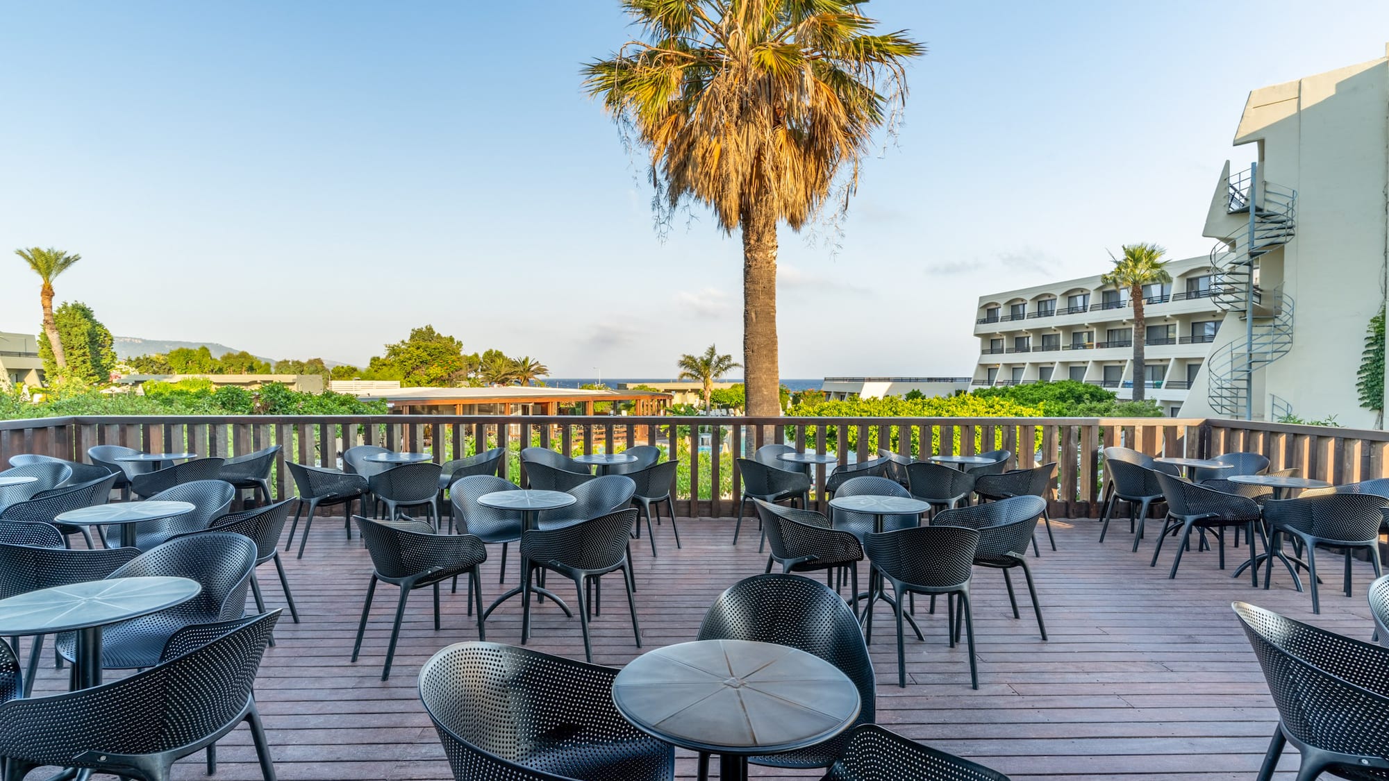Sunny hotel patio with black outdoor seating, palm tree, and distant sea view.