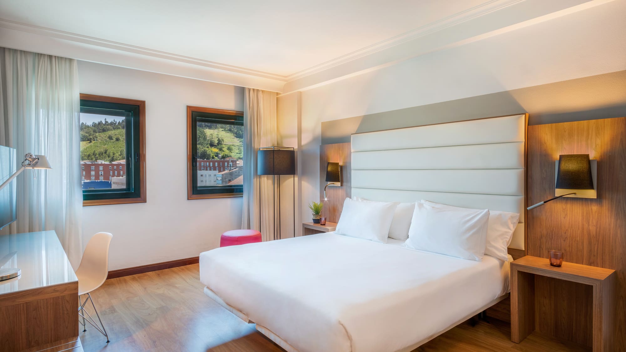 Modern hotel room with king bed, wooden floor, desk, and hillside city view.