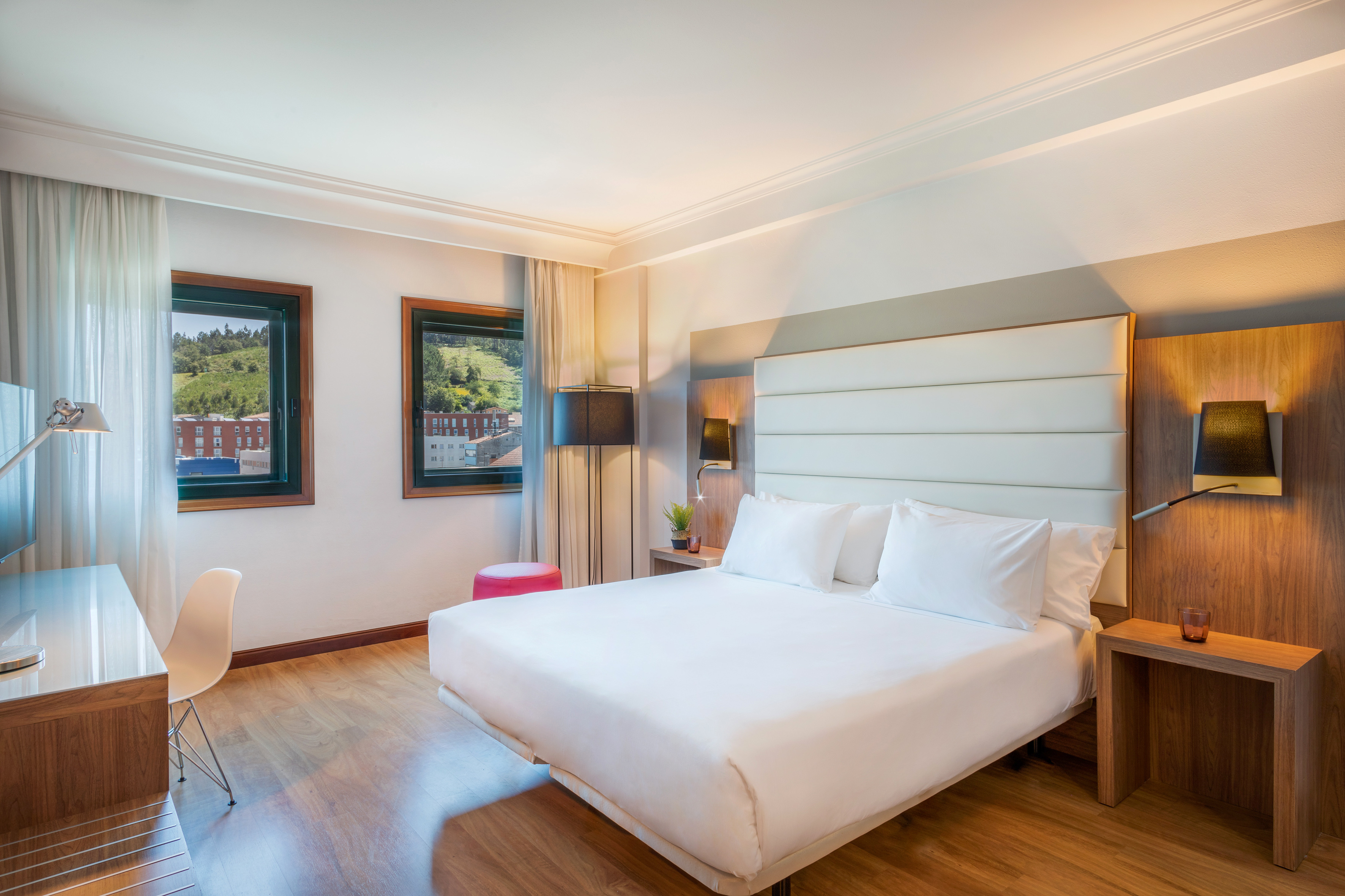 Modern hotel room with king bed, wooden floor, desk, and hillside city view.