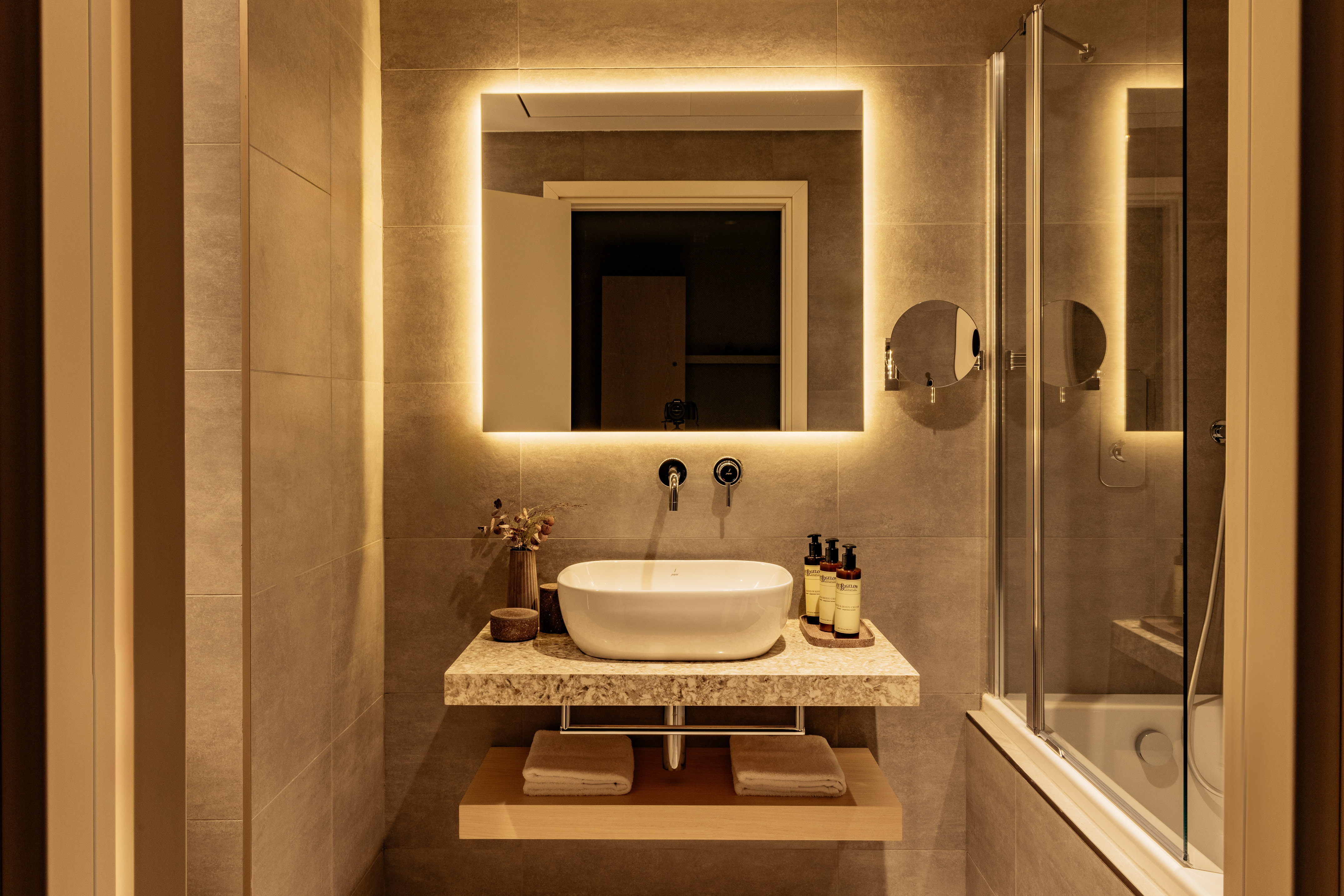 Modern hotel bathroom with vessel sink vanity, backlit mirror, and shower area.