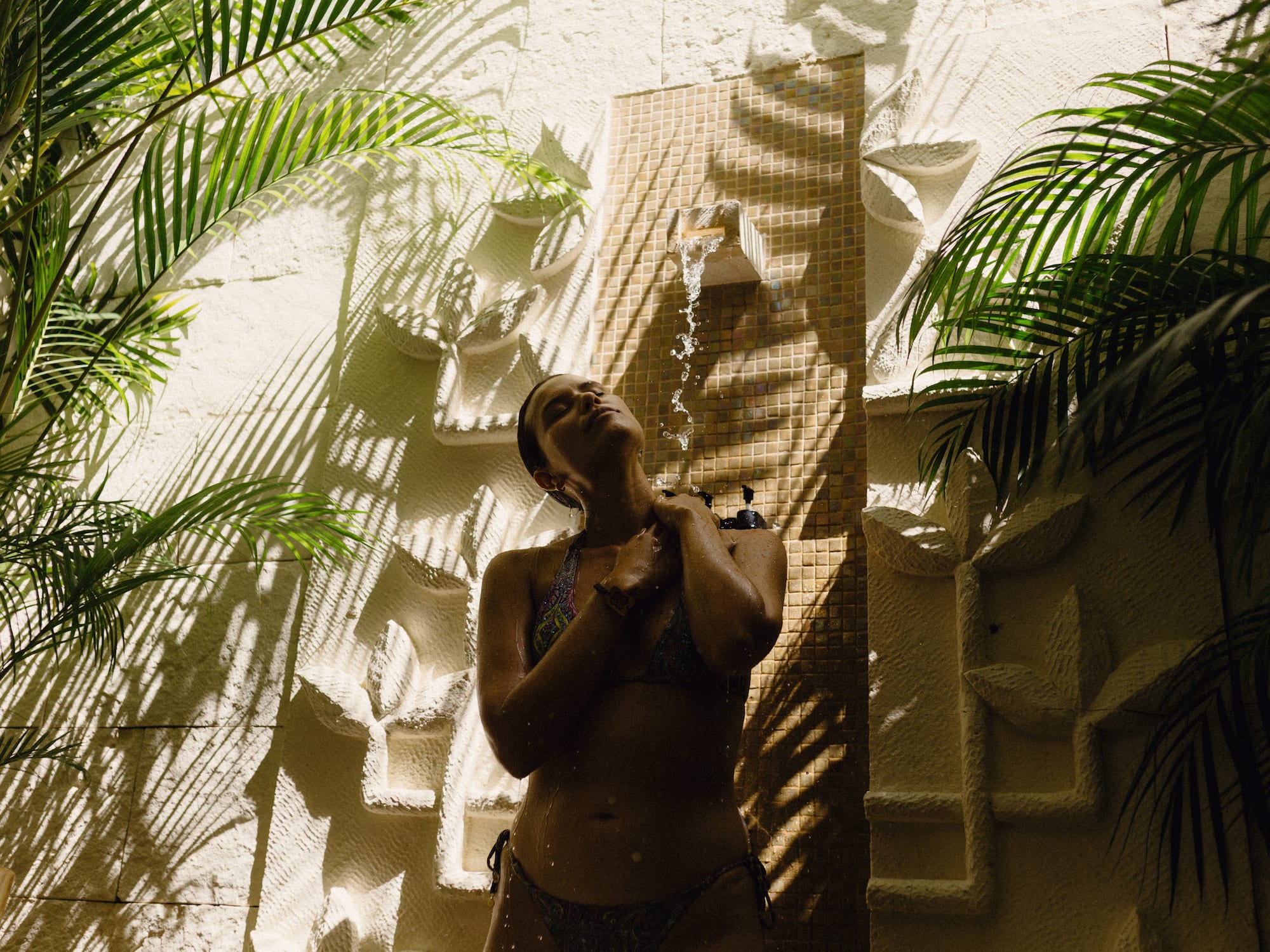 Woman in bikini showering outdoors with tropical palm leaves and stone wall.