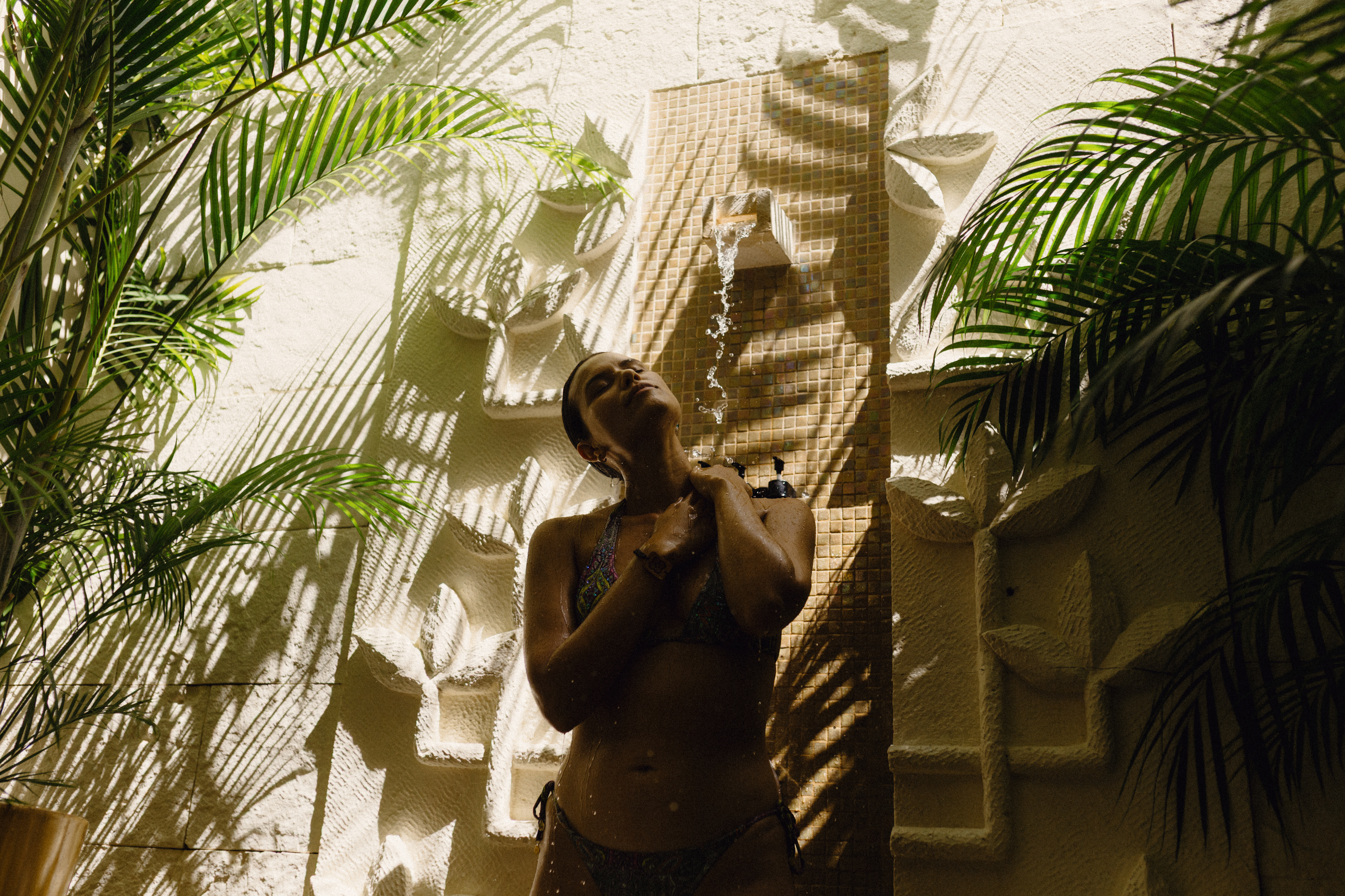 Woman in bikini showering outdoors with tropical palm leaves and stone wall.