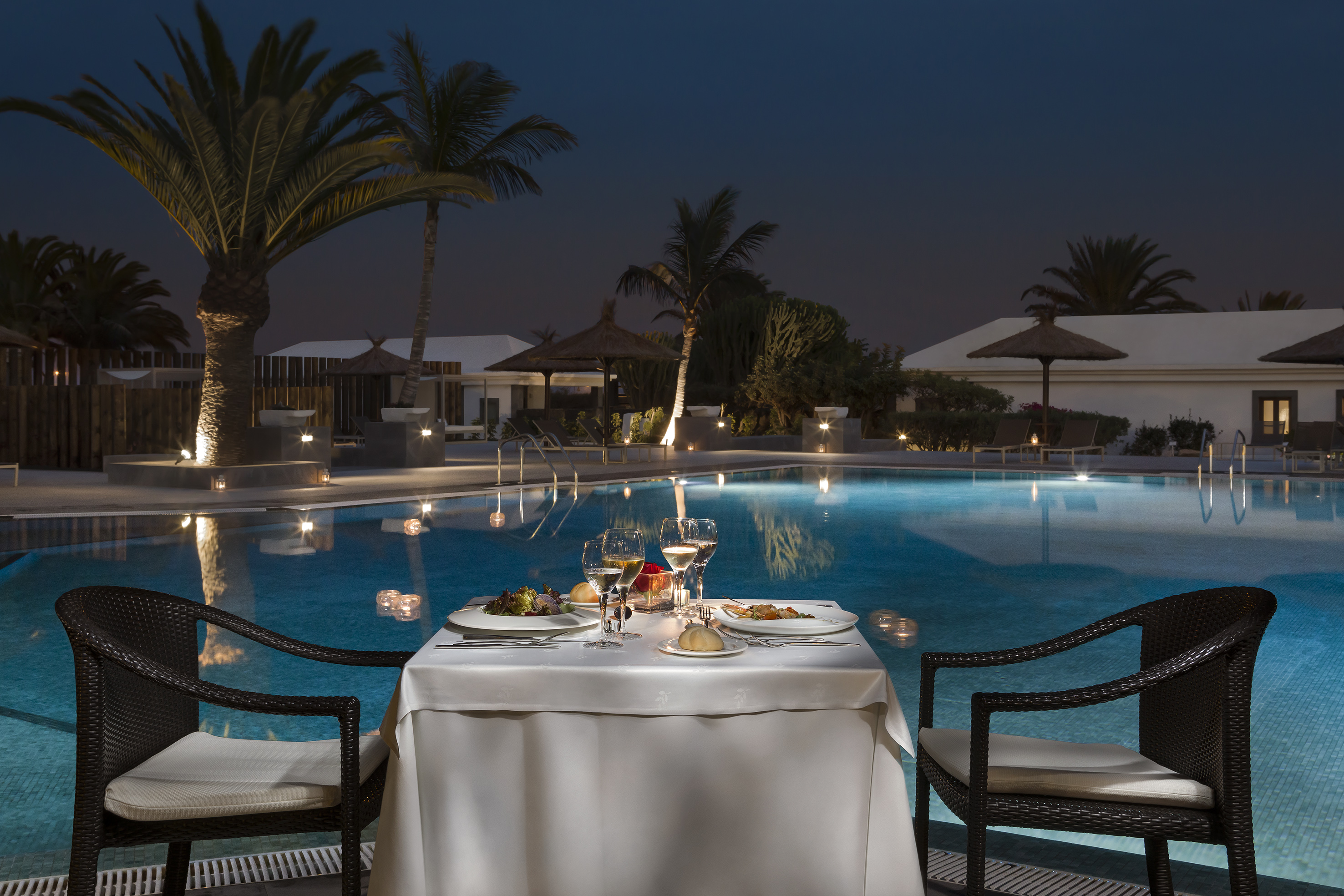 Romantic dinner at a resort pool at night, with palm trees and candlelight.