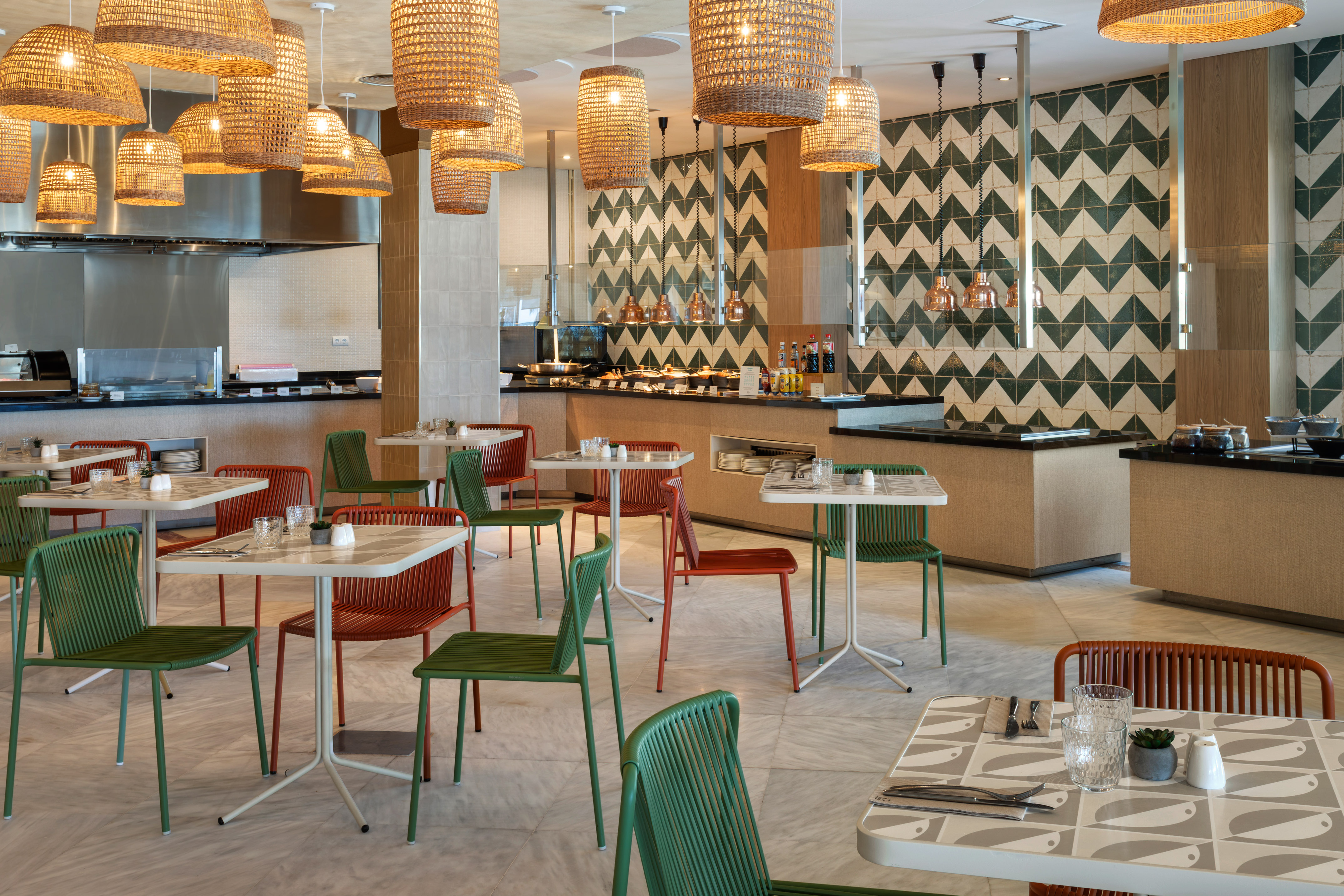 Modern restaurant buffet with green and orange chairs, wicker lamps, tiled walls.