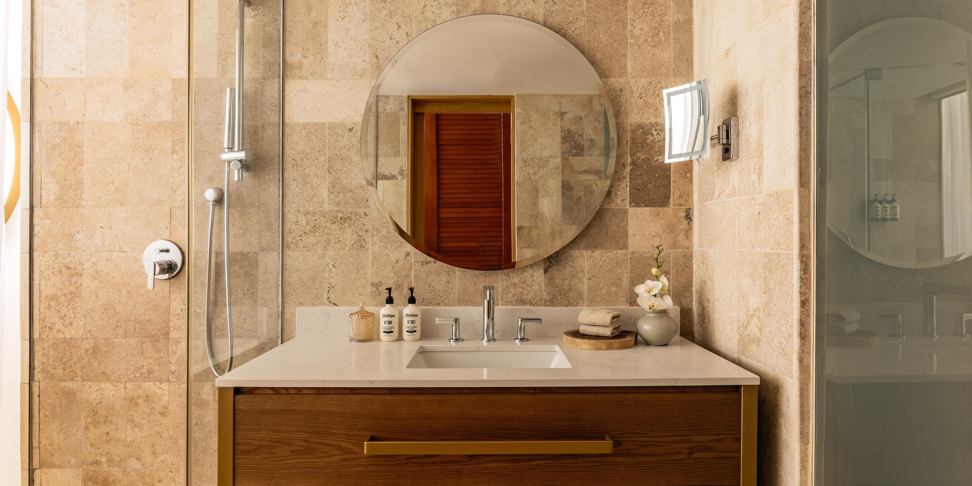 Modern bathroom with wood vanity, round mirror, white counter, and tiled shower.