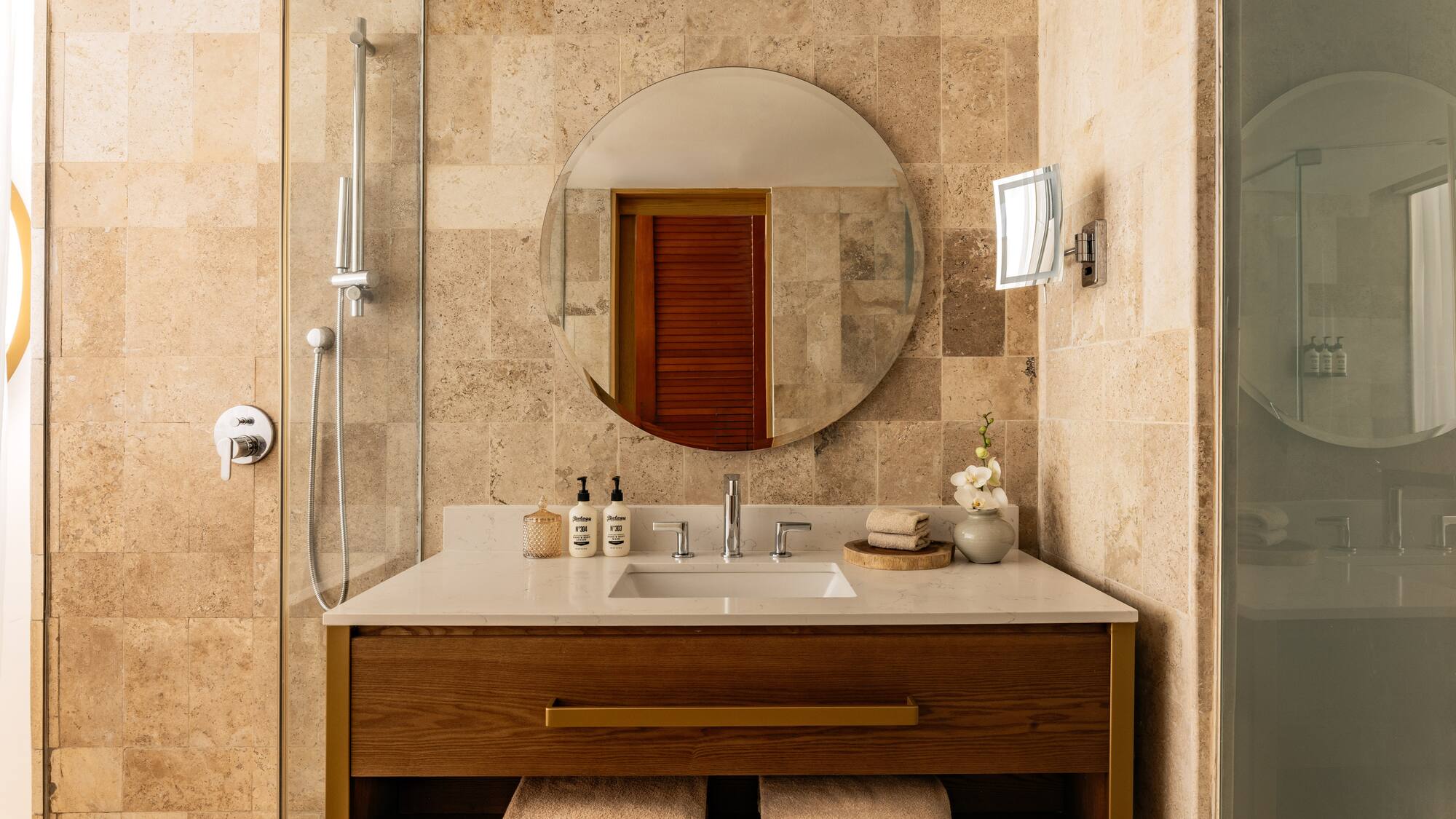 Modern bathroom with wood vanity, round mirror, white counter, and tiled shower.