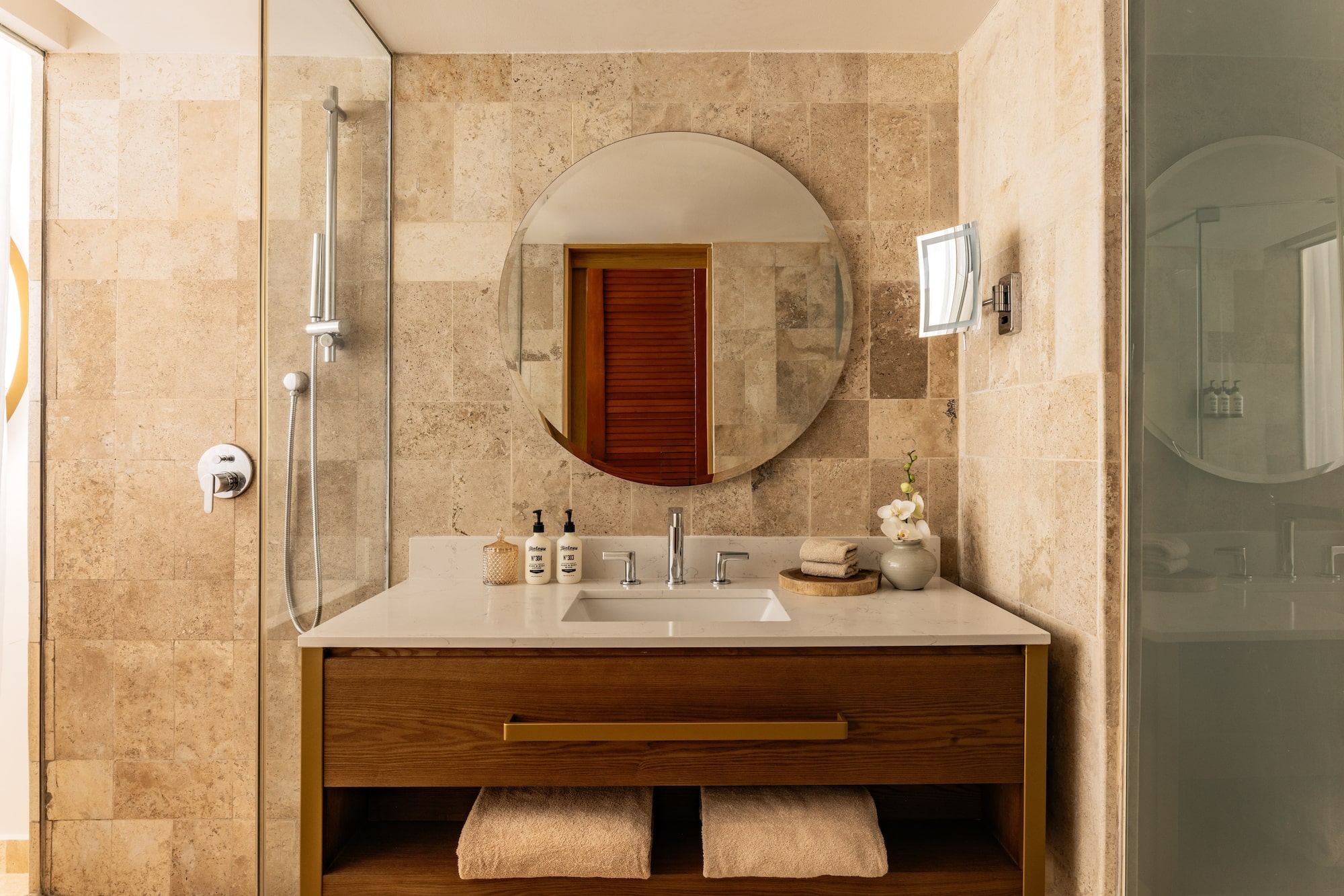 Modern bathroom with wood vanity, round mirror, white counter, and tiled shower.