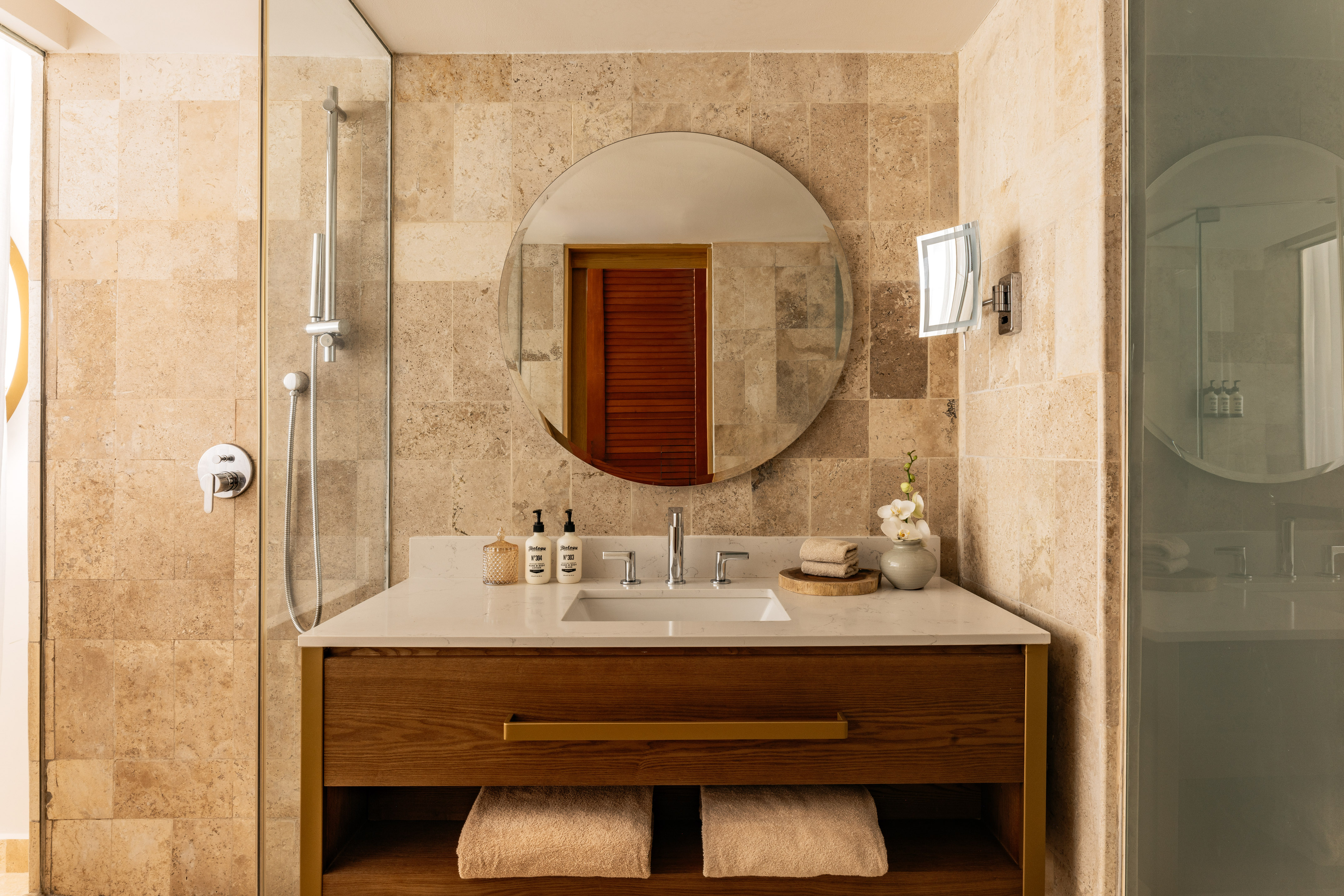 Modern bathroom with wood vanity, round mirror, white counter, and tiled shower.