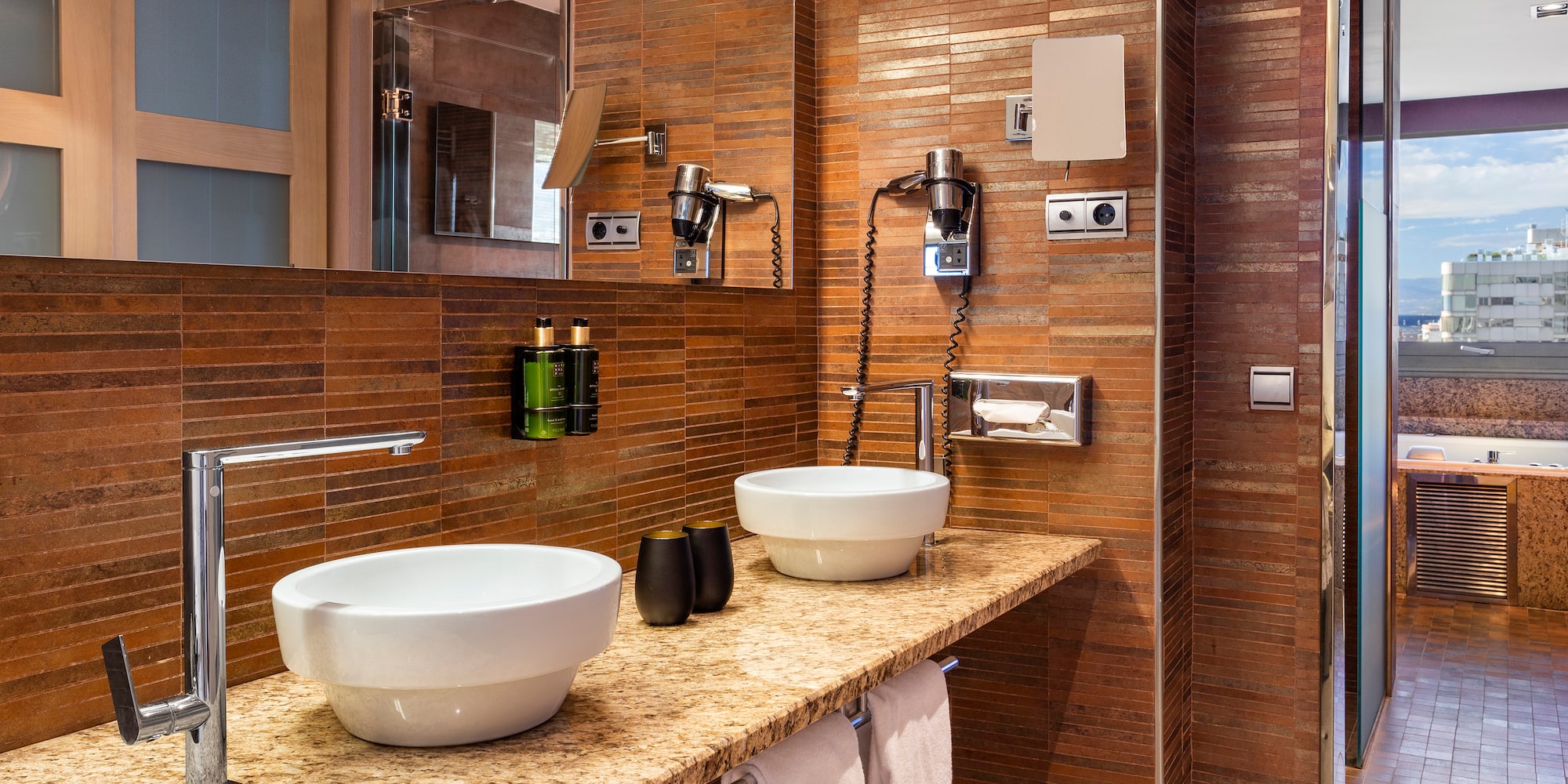 Baño de hotel moderno con dos lavabos, encimera de granito y azulejos terracota.