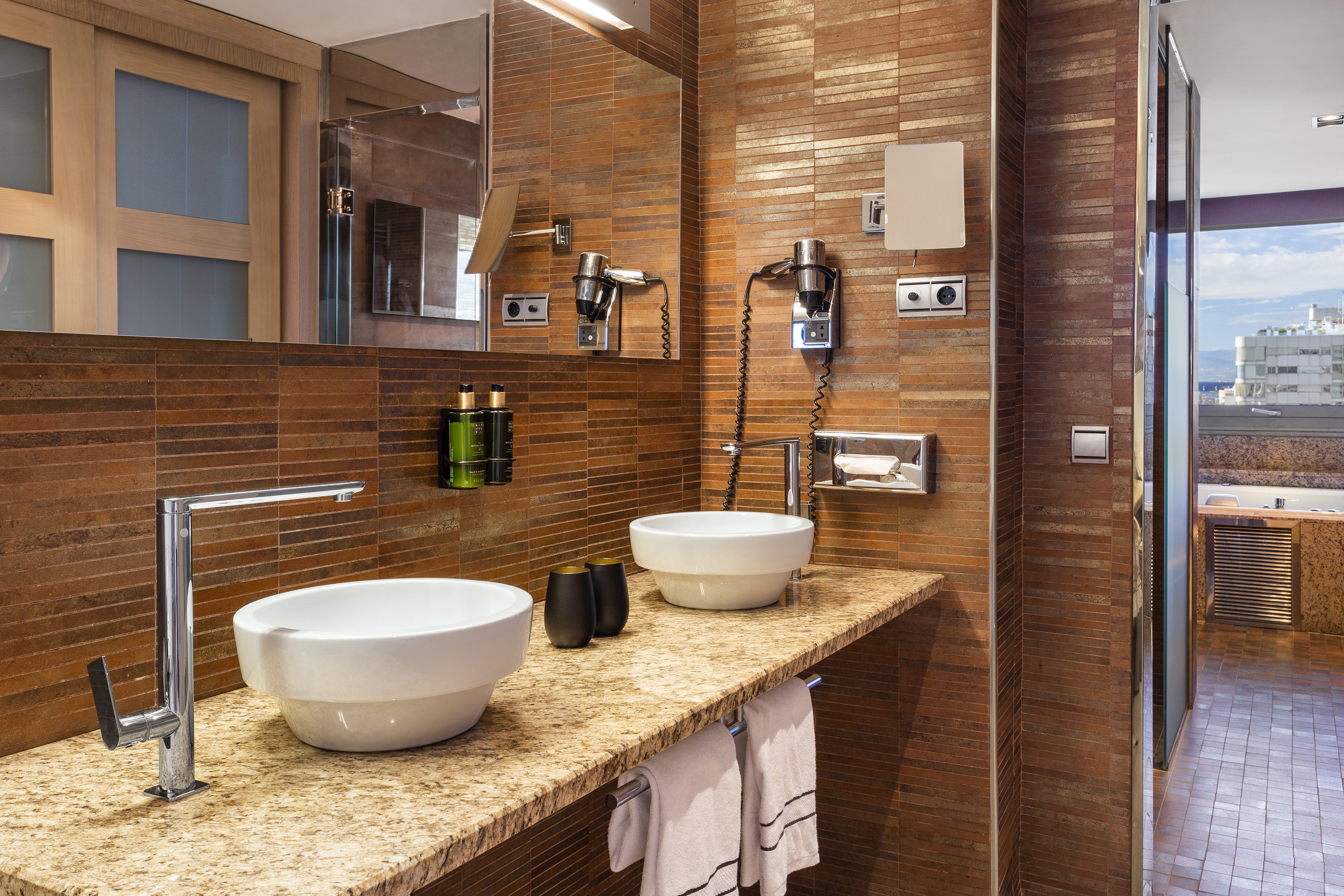 Baño de hotel moderno con dos lavabos, encimera de granito y azulejos terracota.