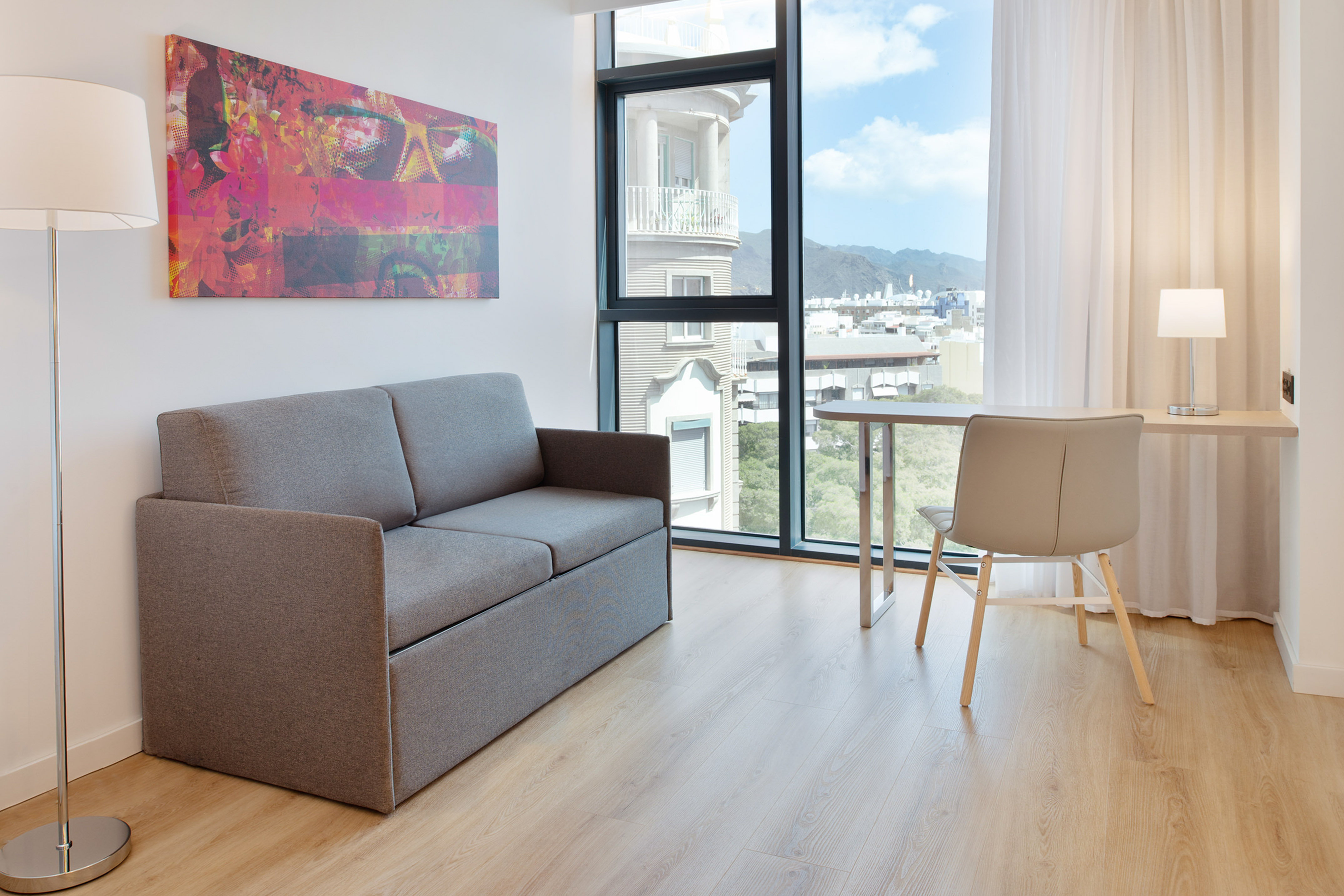 Modern hotel room with grey sofa, desk, and city view from large window.