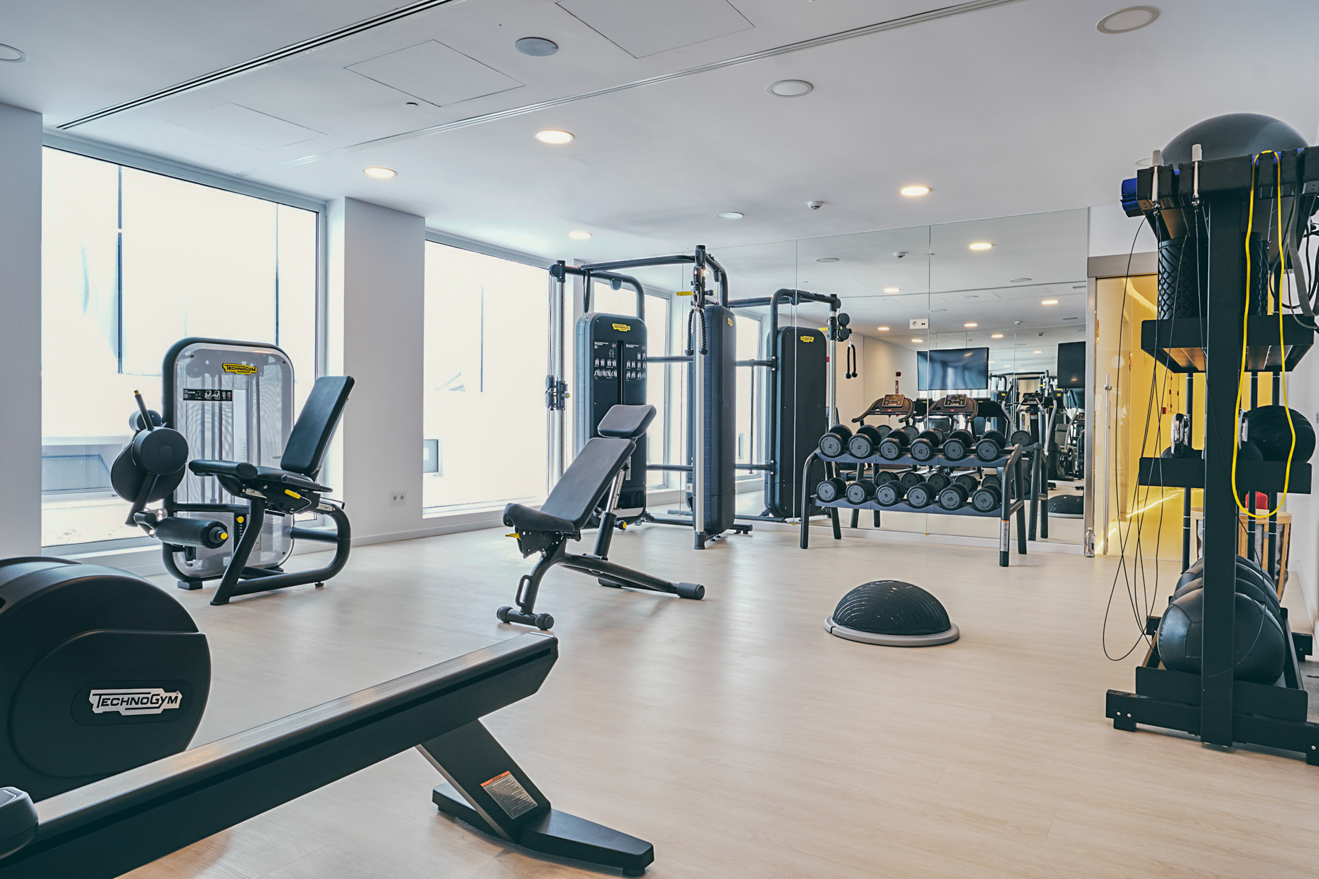 Modern fitness center with Technogym equipment, dumbbell rack, and large windows.