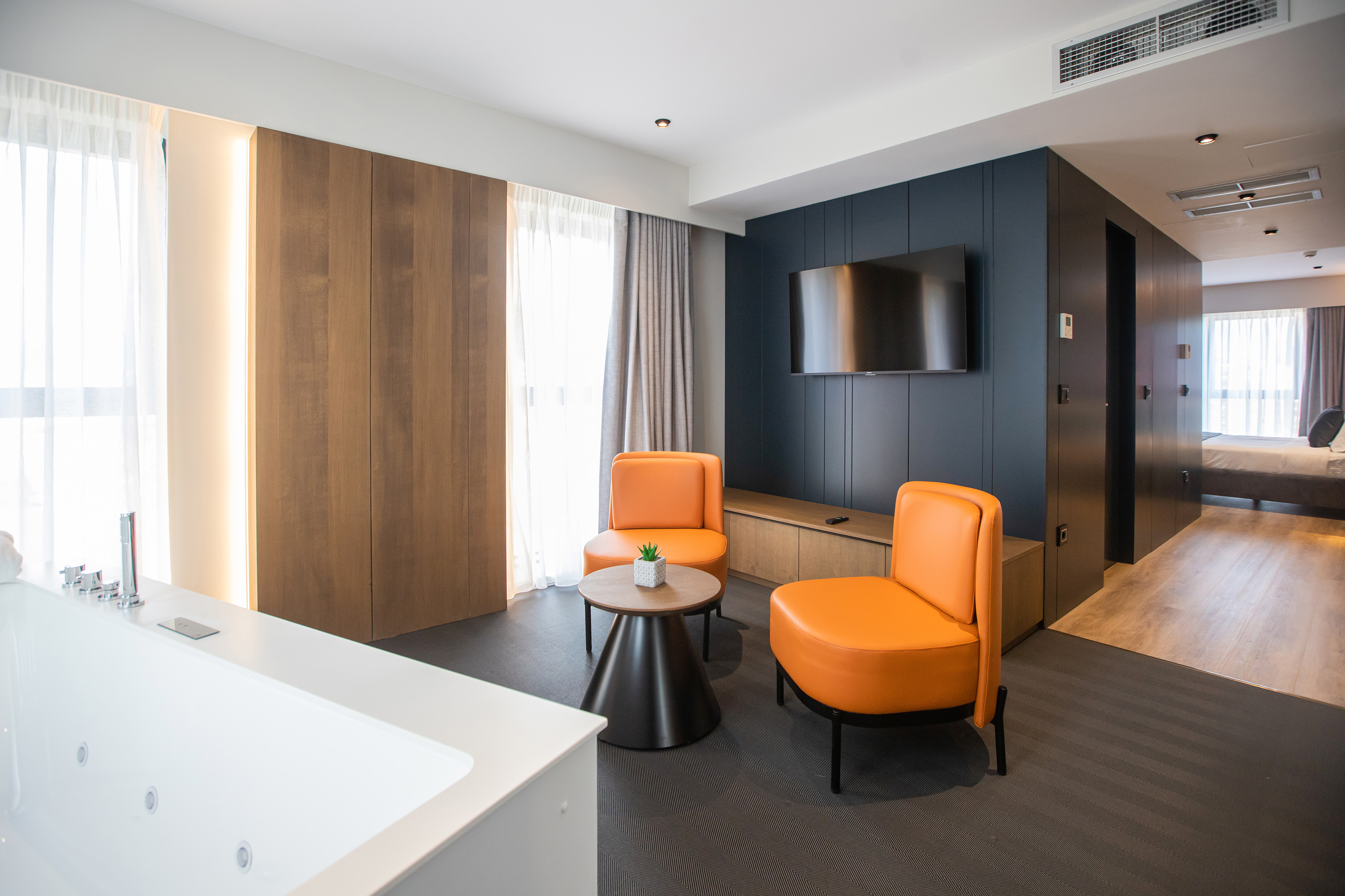 Modern hotel suite with orange armchairs, wall TV, and jacuzzi tub.