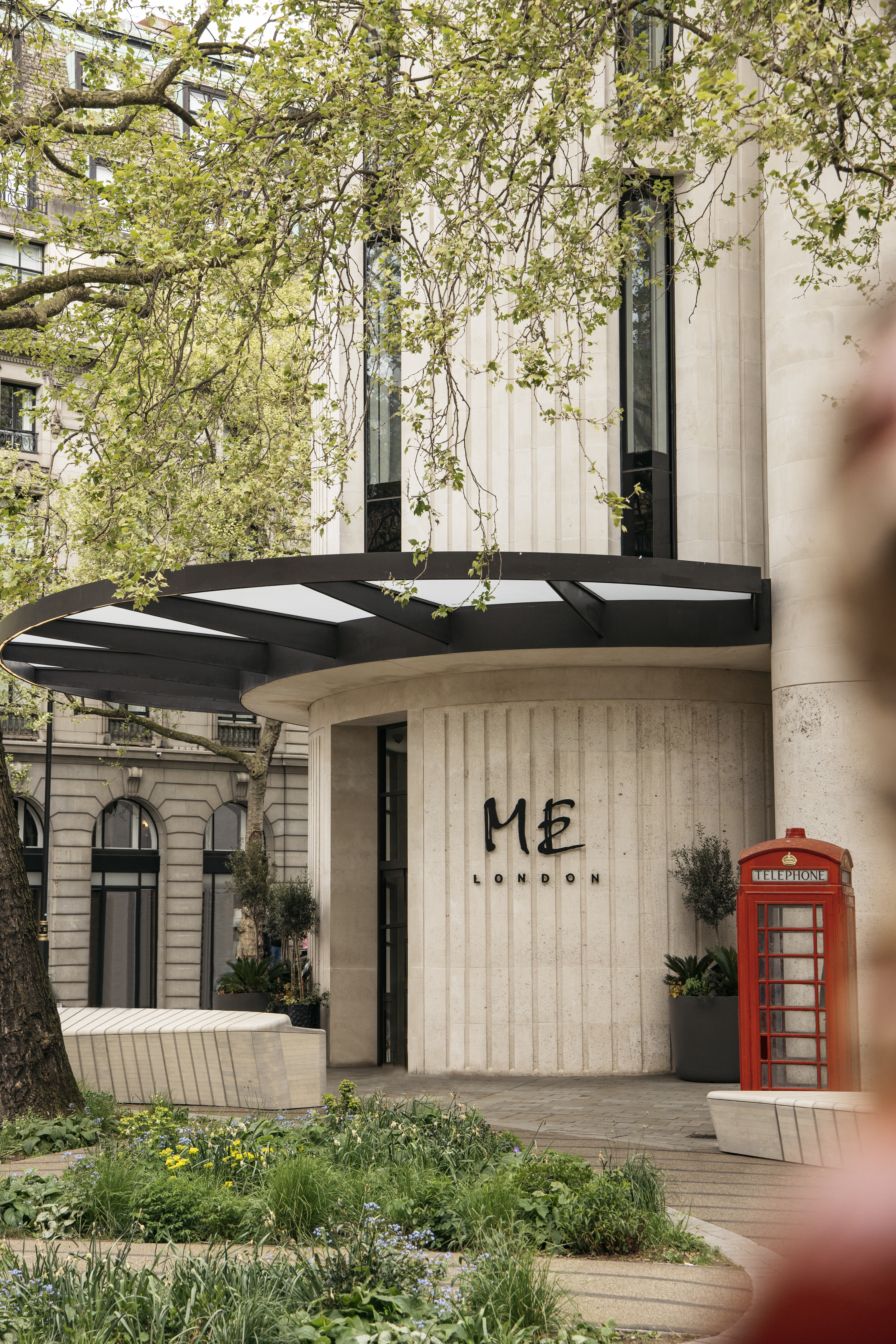 Modern ME London hotel facade, red telephone box, leafy spring trees.