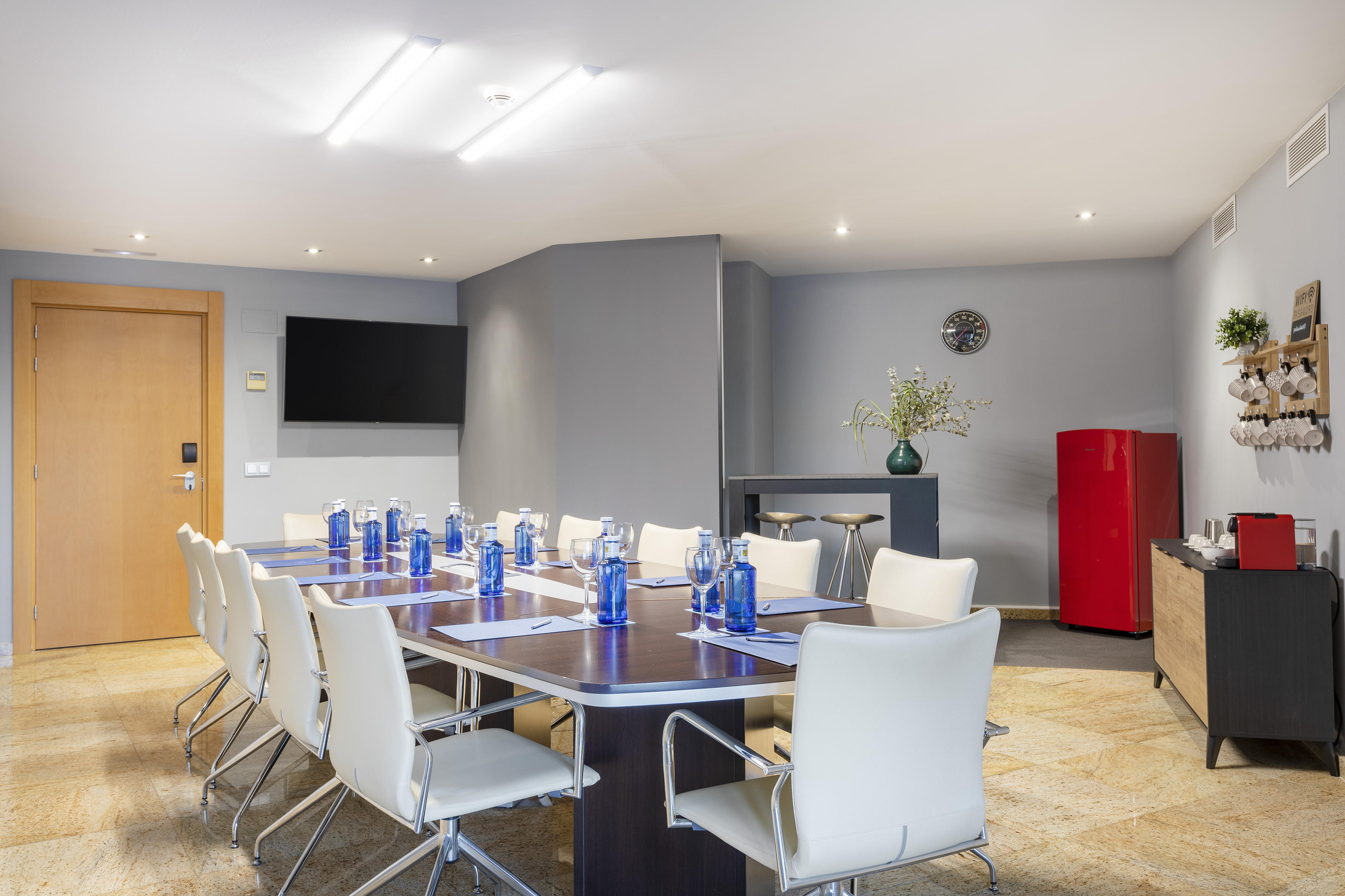 Modern meeting room with conference table, white chairs, TV, and coffee station.