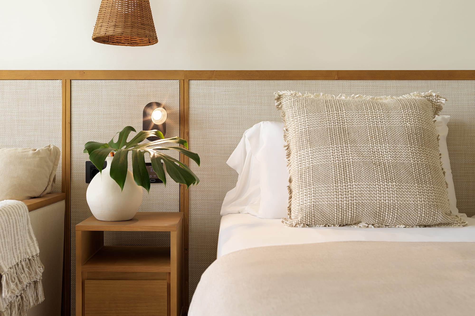 Bedroom with cane headboard, wood nightstand, plant, and neutral bedding.