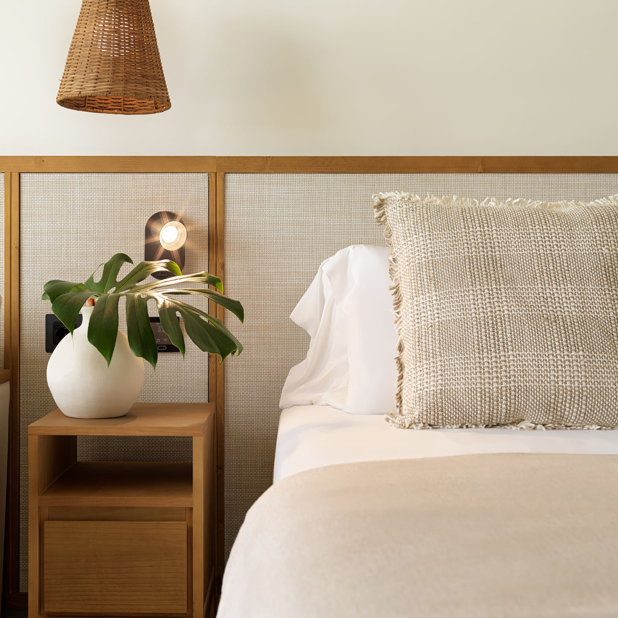 Bedroom with cane headboard, wood nightstand, plant, and neutral bedding.
