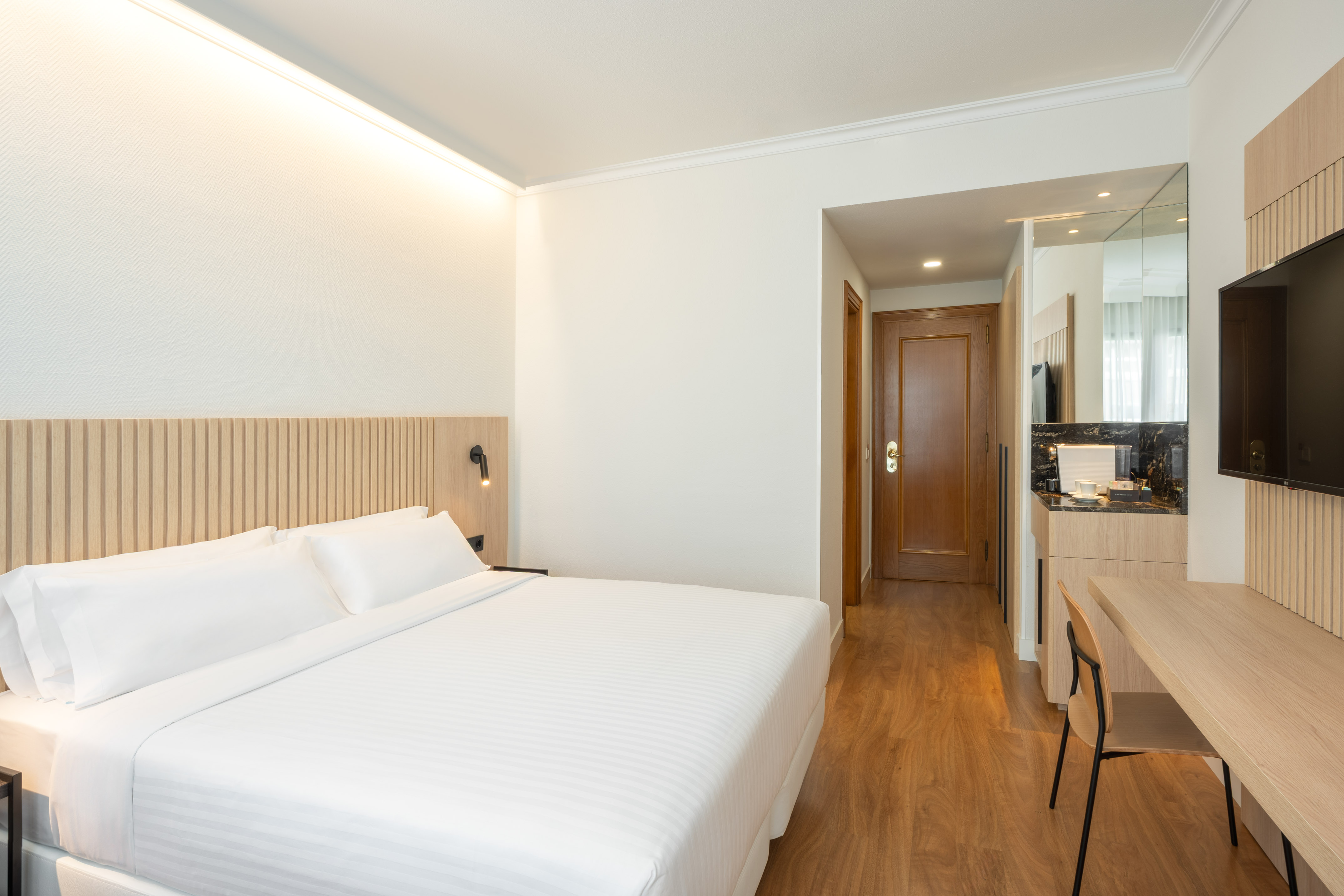 Modern hotel room with white bed, wooden headboard, desk, and TV.