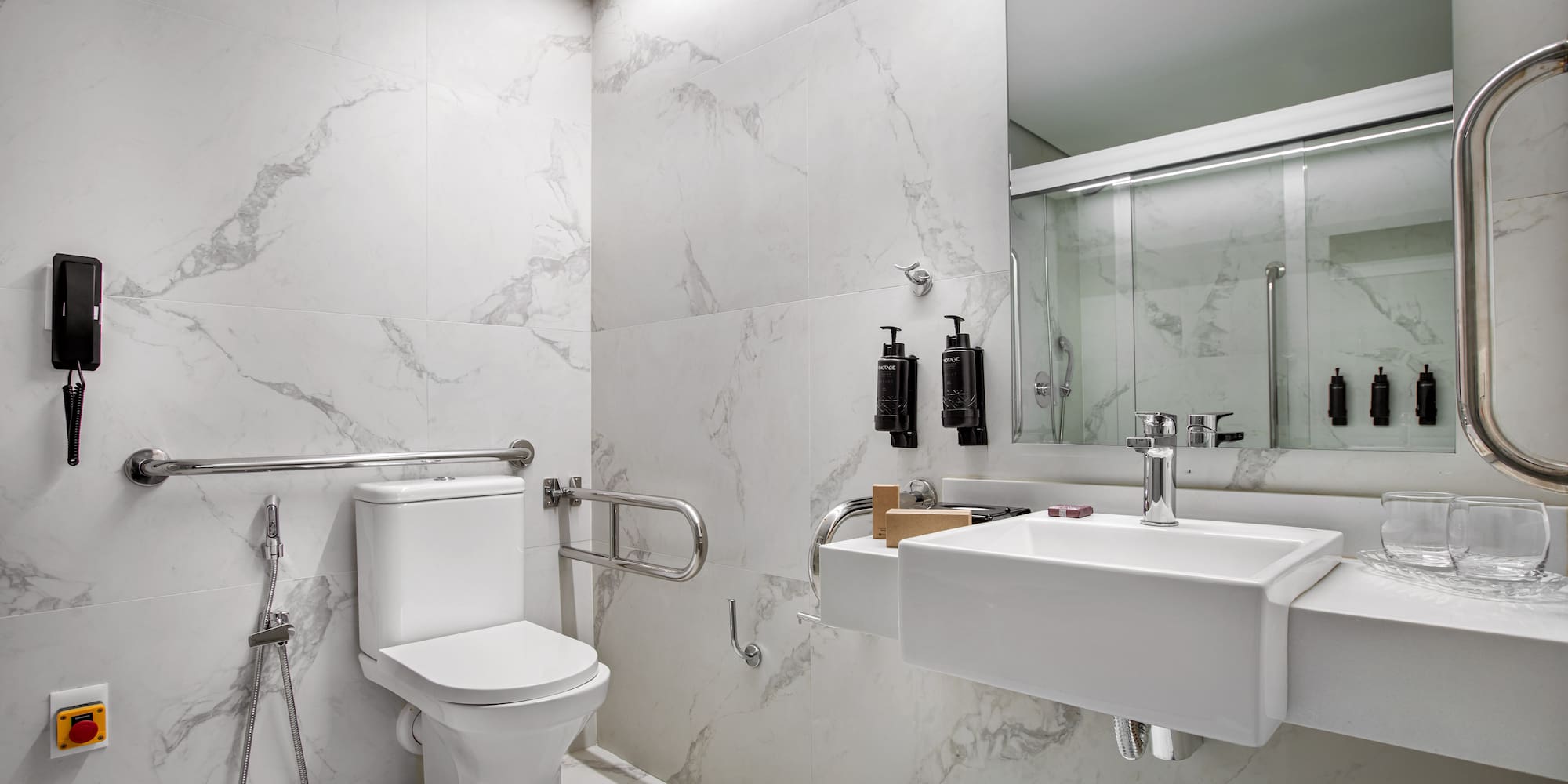 Accessible modern bathroom with white marble, toilet, sink, grab bars, and shower.