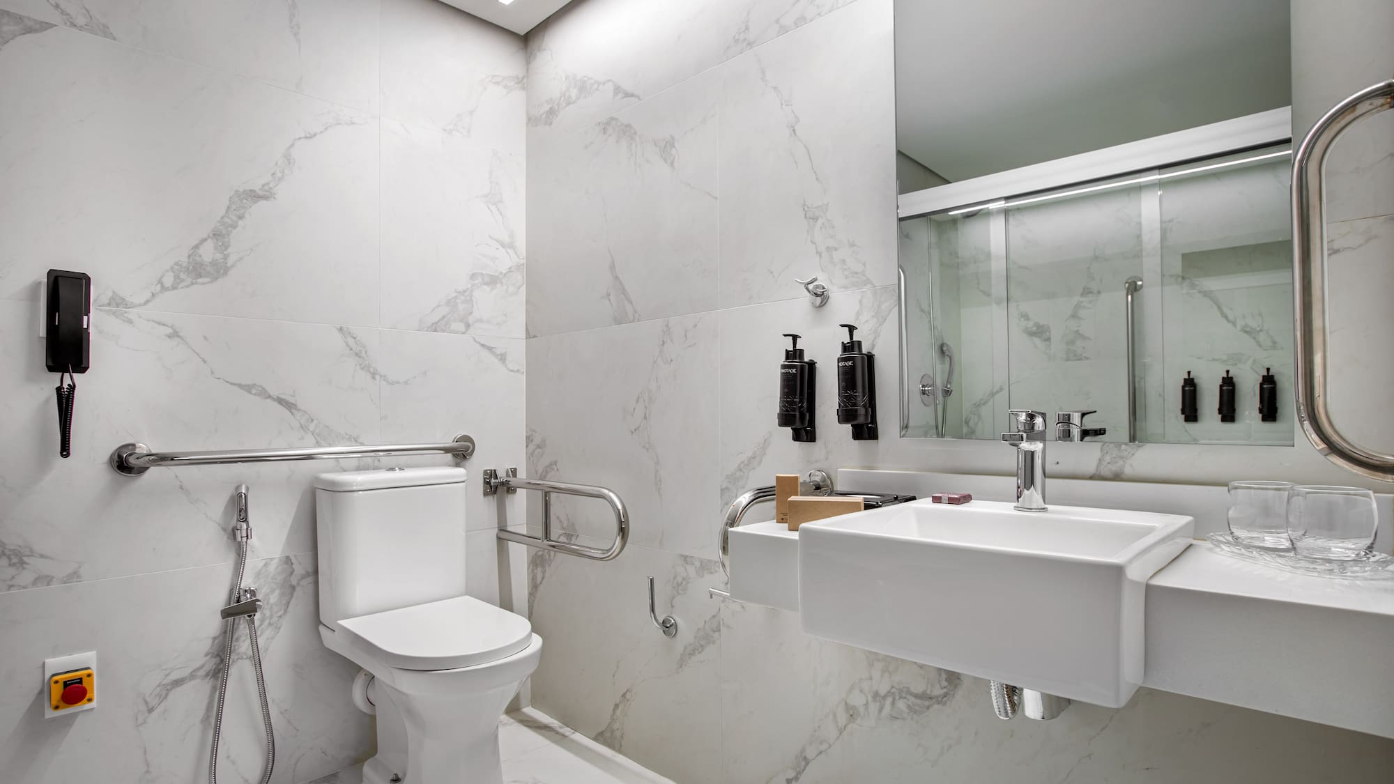 Accessible modern bathroom with white marble, toilet, sink, grab bars, and shower.