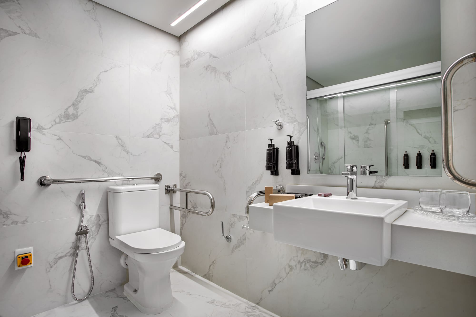 Accessible modern bathroom with white marble, toilet, sink, grab bars, and shower.