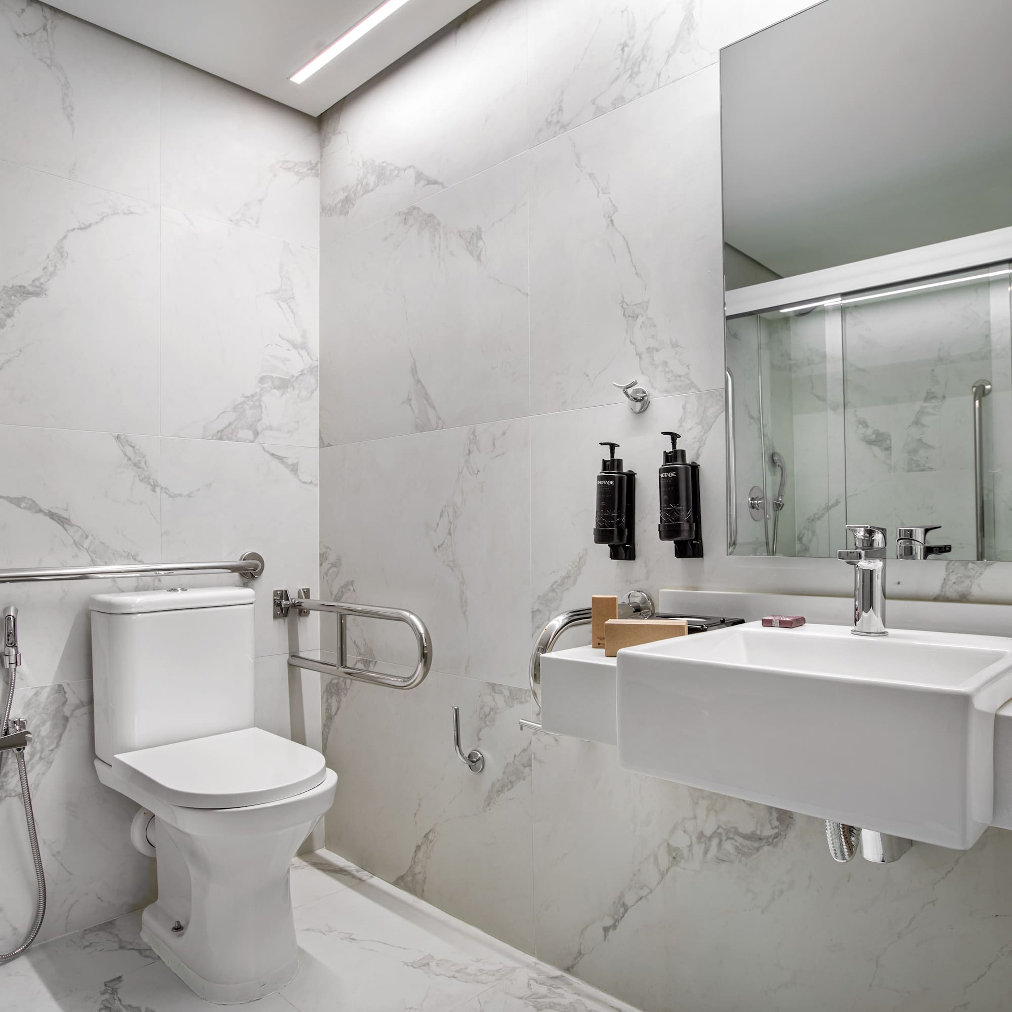 Accessible modern bathroom with white marble, toilet, sink, grab bars, and shower.