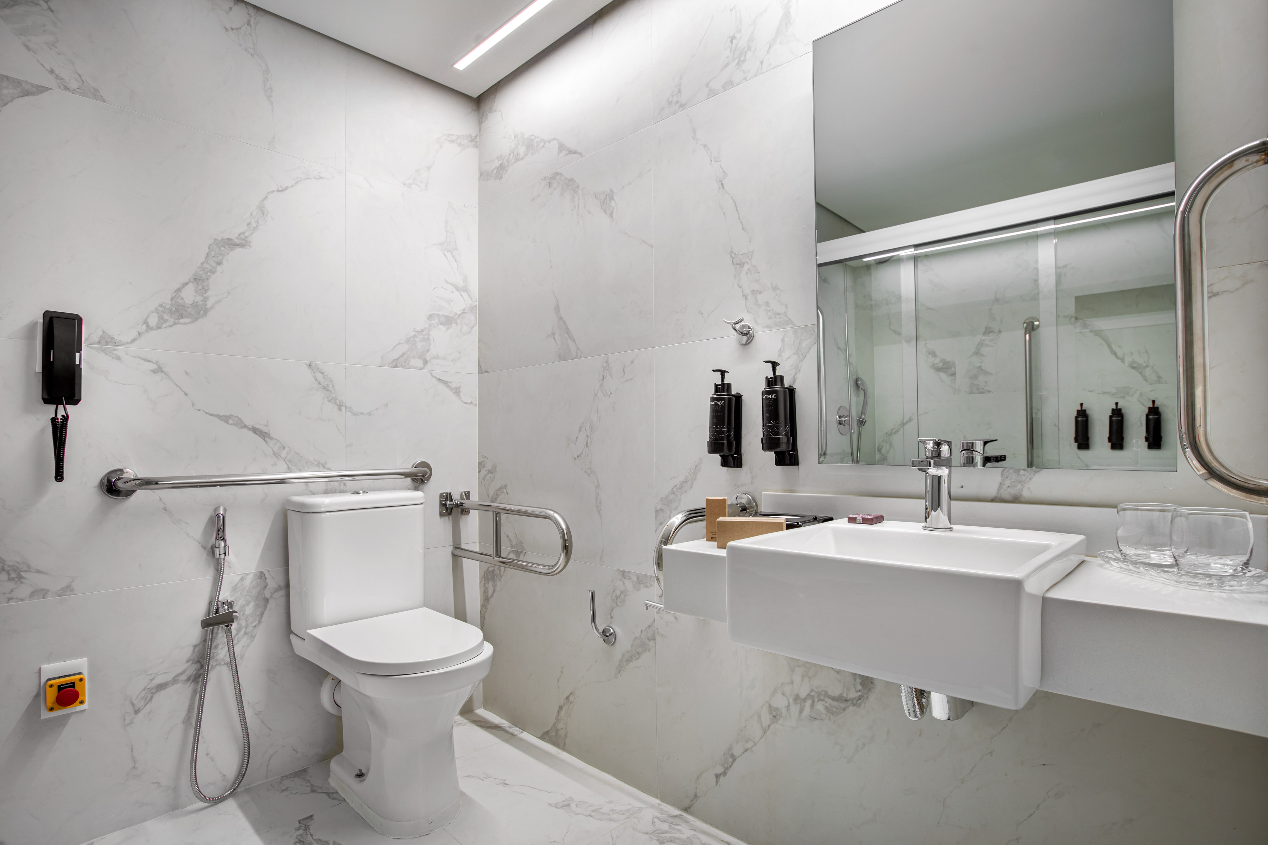 Accessible modern bathroom with white marble, toilet, sink, grab bars, and shower.