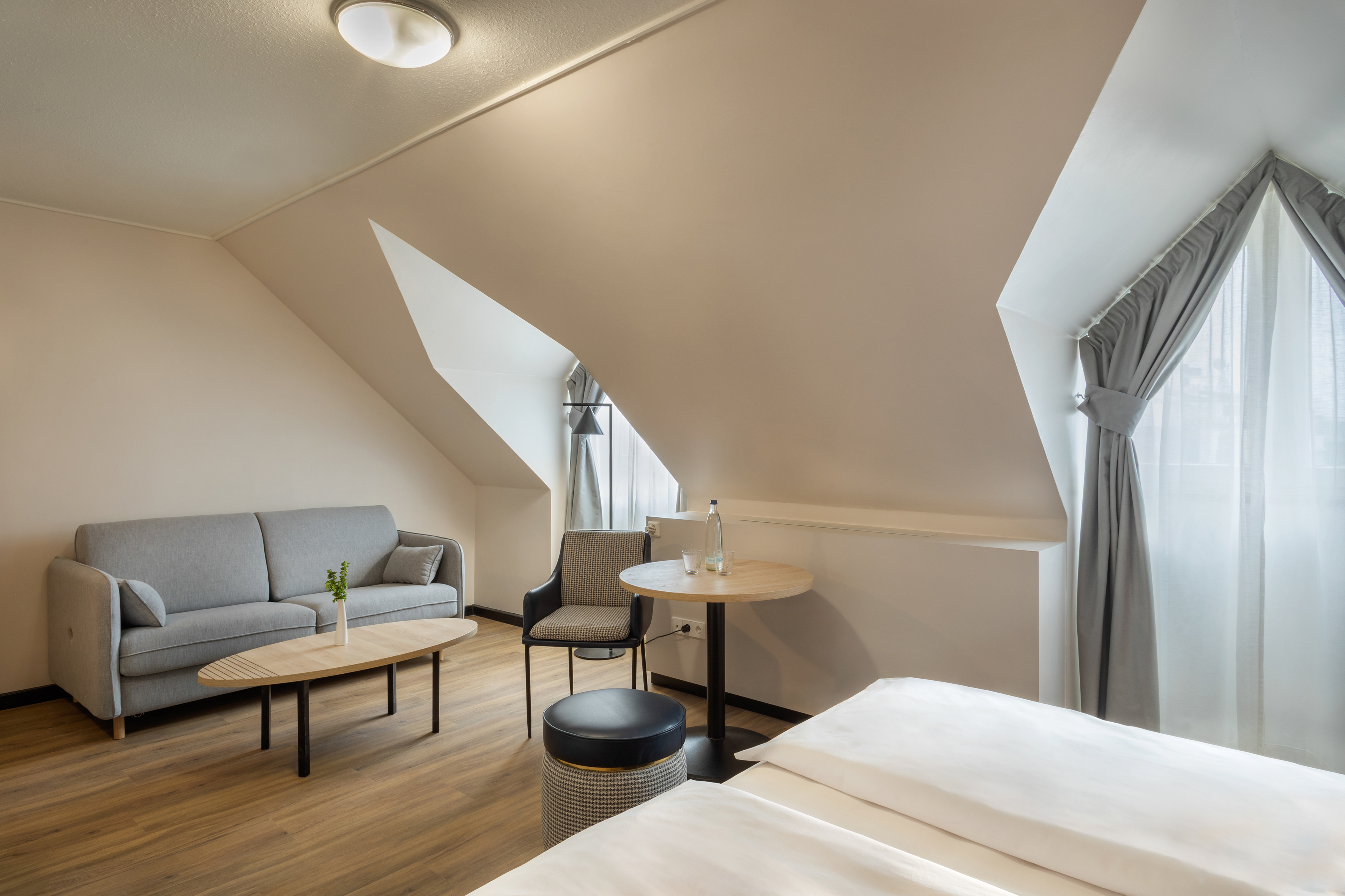 Modern hotel suite with sloped ceiling, gray sofa, table, and bed.