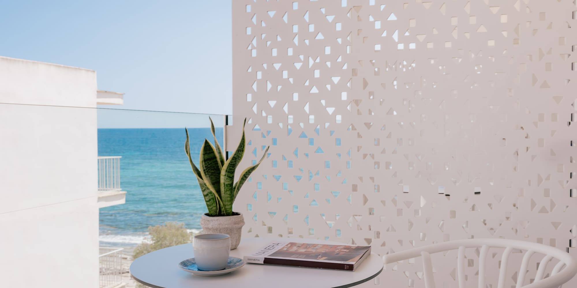 Balcony with sea view, coffee, plant, magazine on white table, patterned wall.