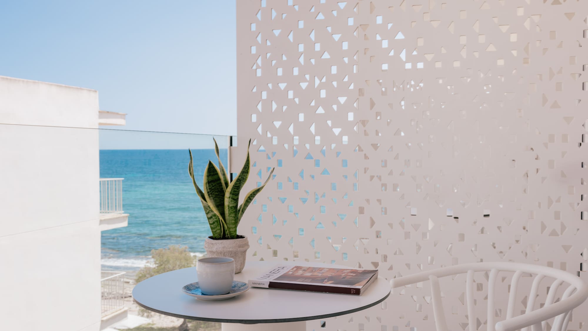 Balcony with sea view, coffee, plant, magazine on white table, patterned wall.