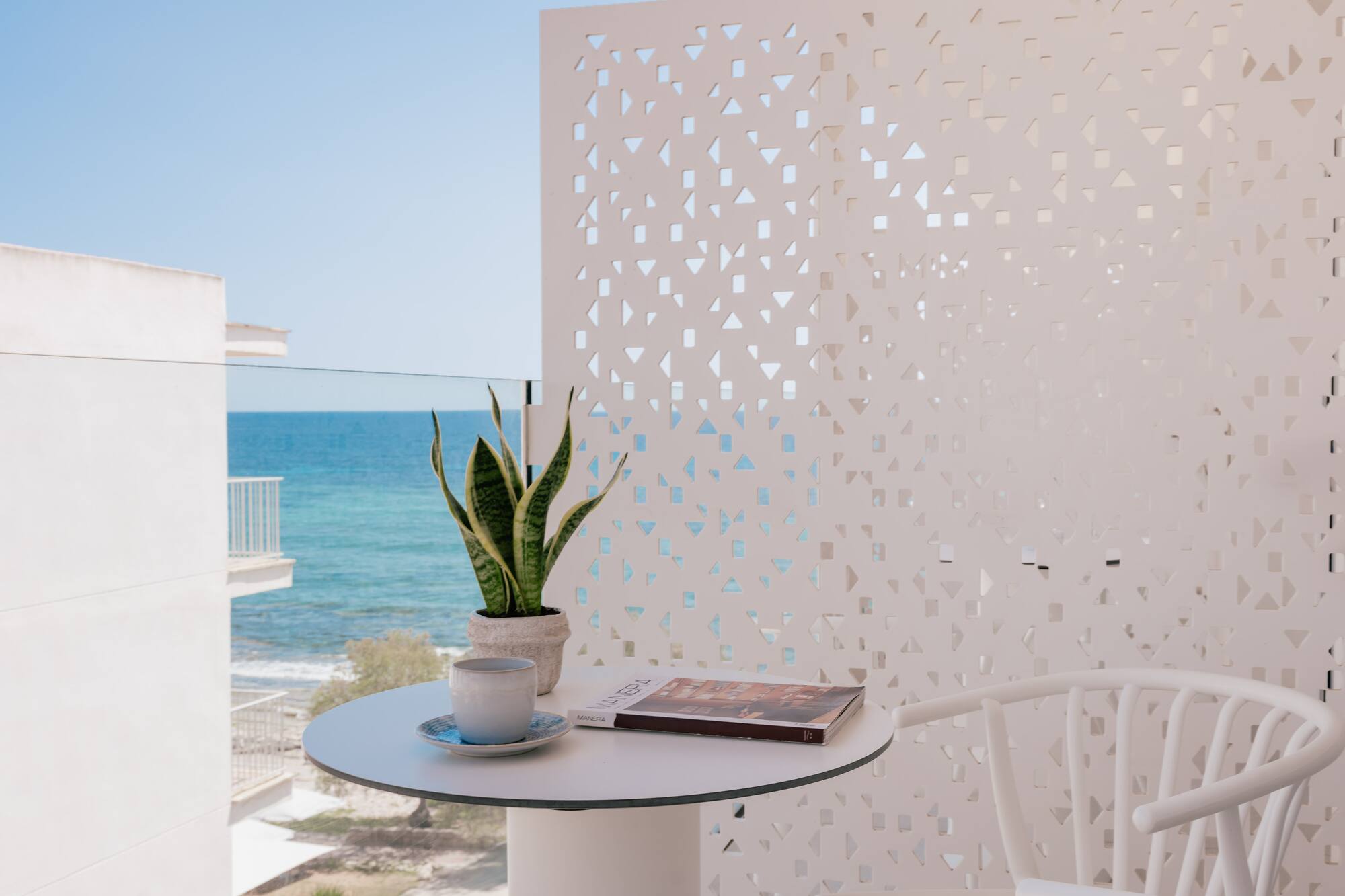 Balcón moderno con vista al mar. Mesa, café, planta, panel decorativo.
