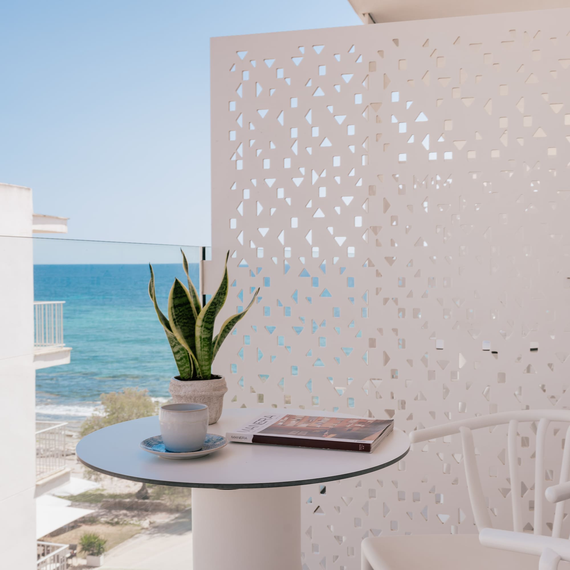 Balcony with sea view, coffee, plant, magazine on white table, patterned wall.
