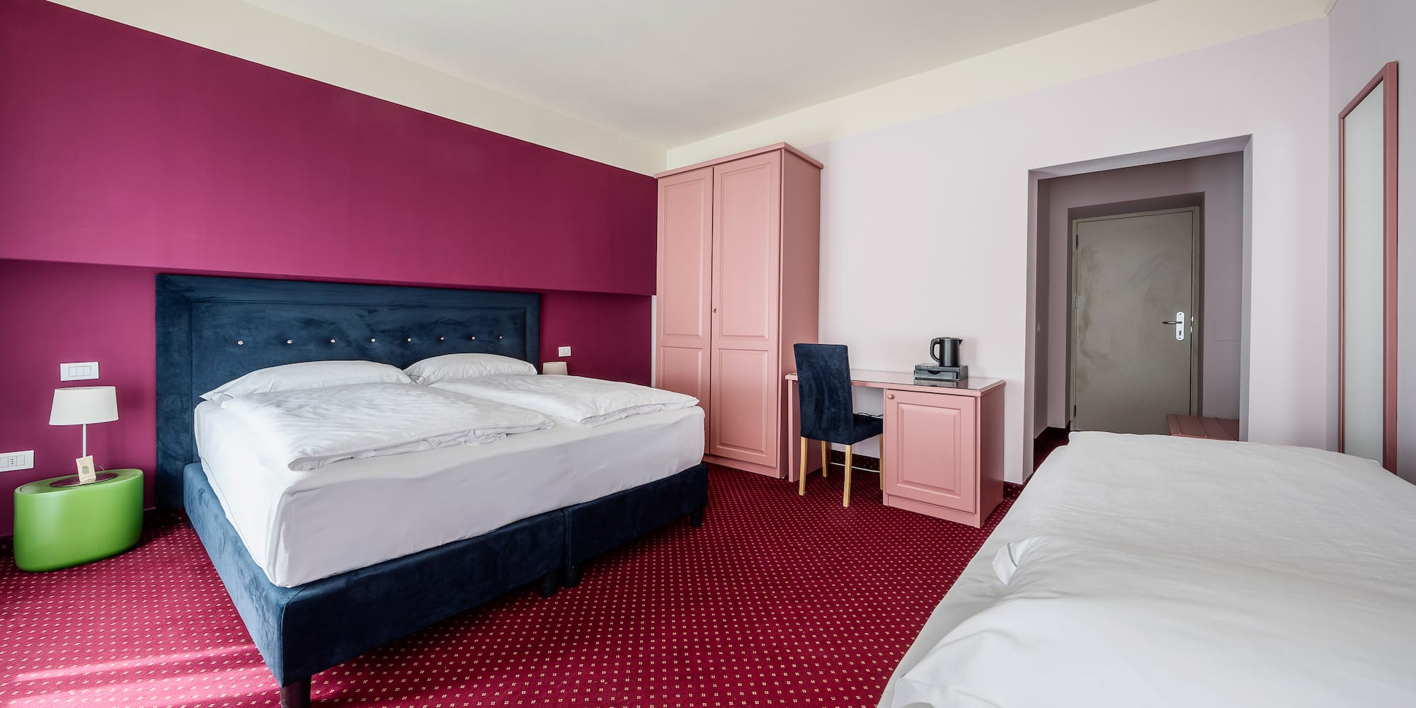 Hotel room with magenta walls, blue bed, red carpet, pink furniture, and extra bed.