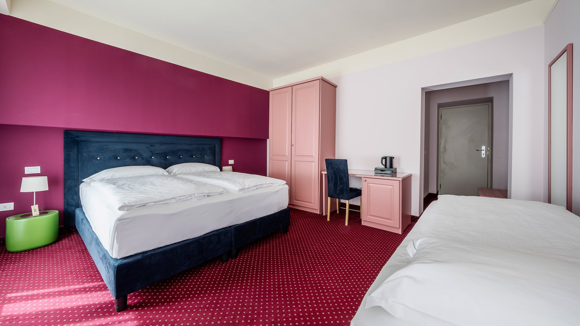 Vibrant hotel room with blue bed, magenta walls, red carpet, and pink desk.