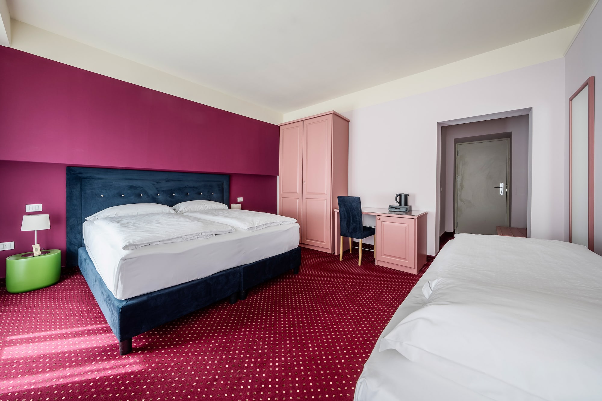 Hotel room with magenta walls, blue bed, red carpet, pink furniture, and extra bed.